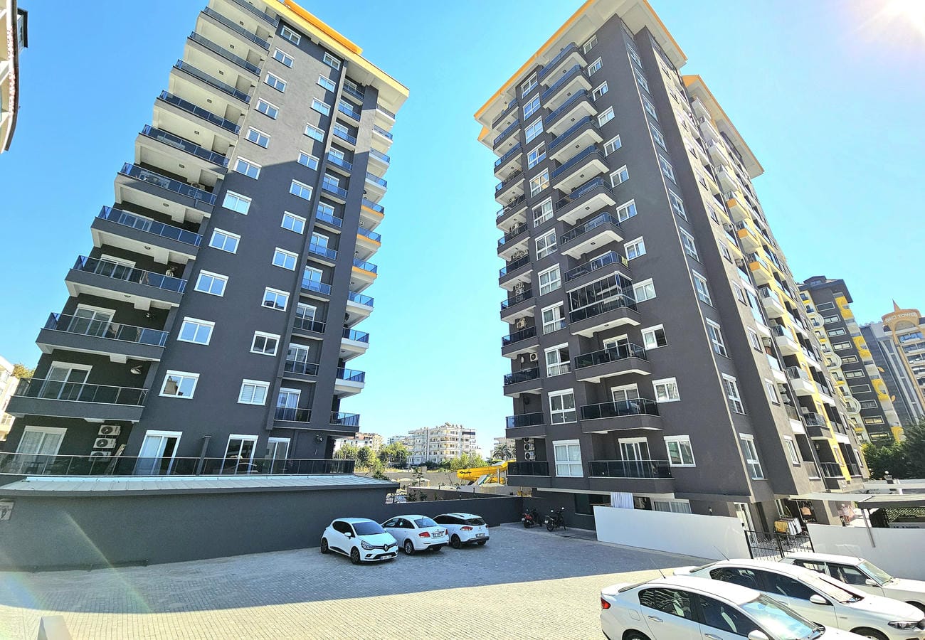 Два жилых корпуса с жёлтыми акцентами, стеклянными балконами и открытой парковкой для квартир 2+1