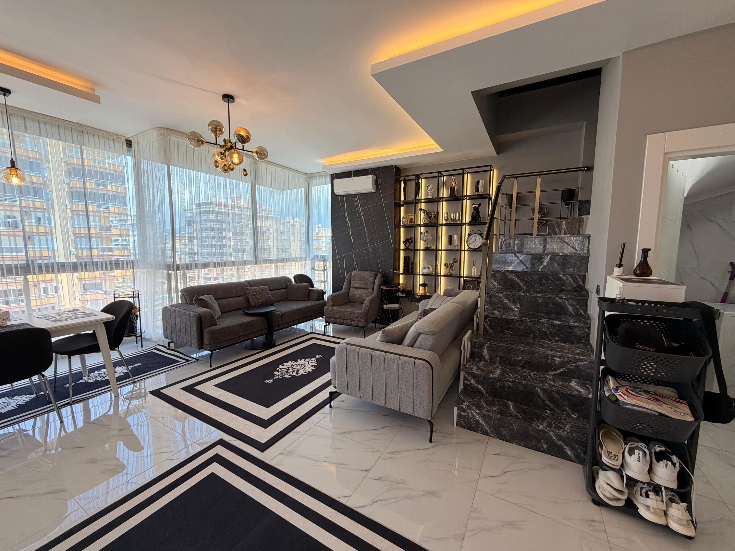 Satılık penthouse mutfak-salonda gri kanepeler, mermer kaplı merdiven ve ışıklı raf