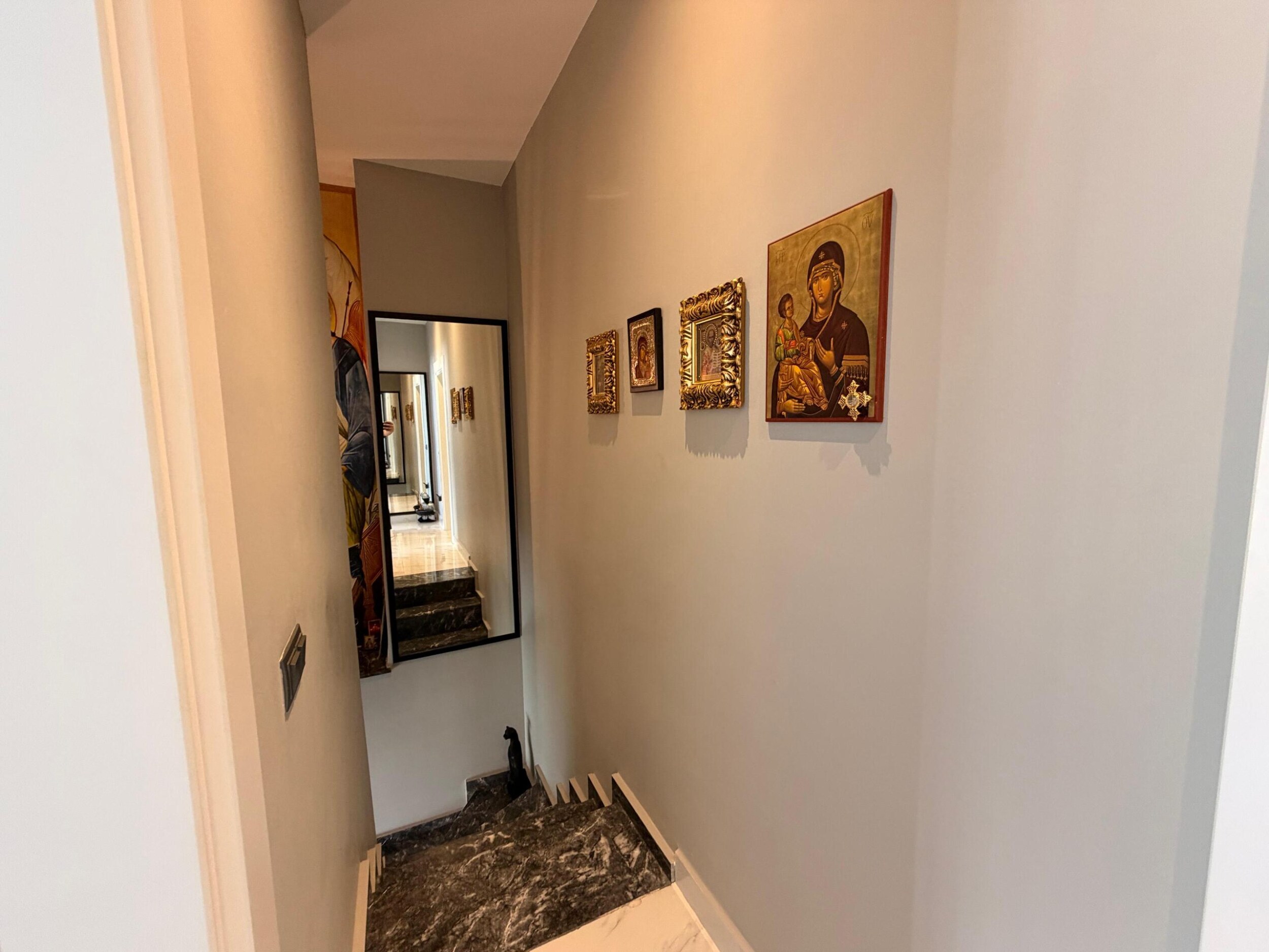 Penthouse merdiveninde koyu mermer basamaklar, çerçeveli ikonalar ve siyah çerçeveli ayna