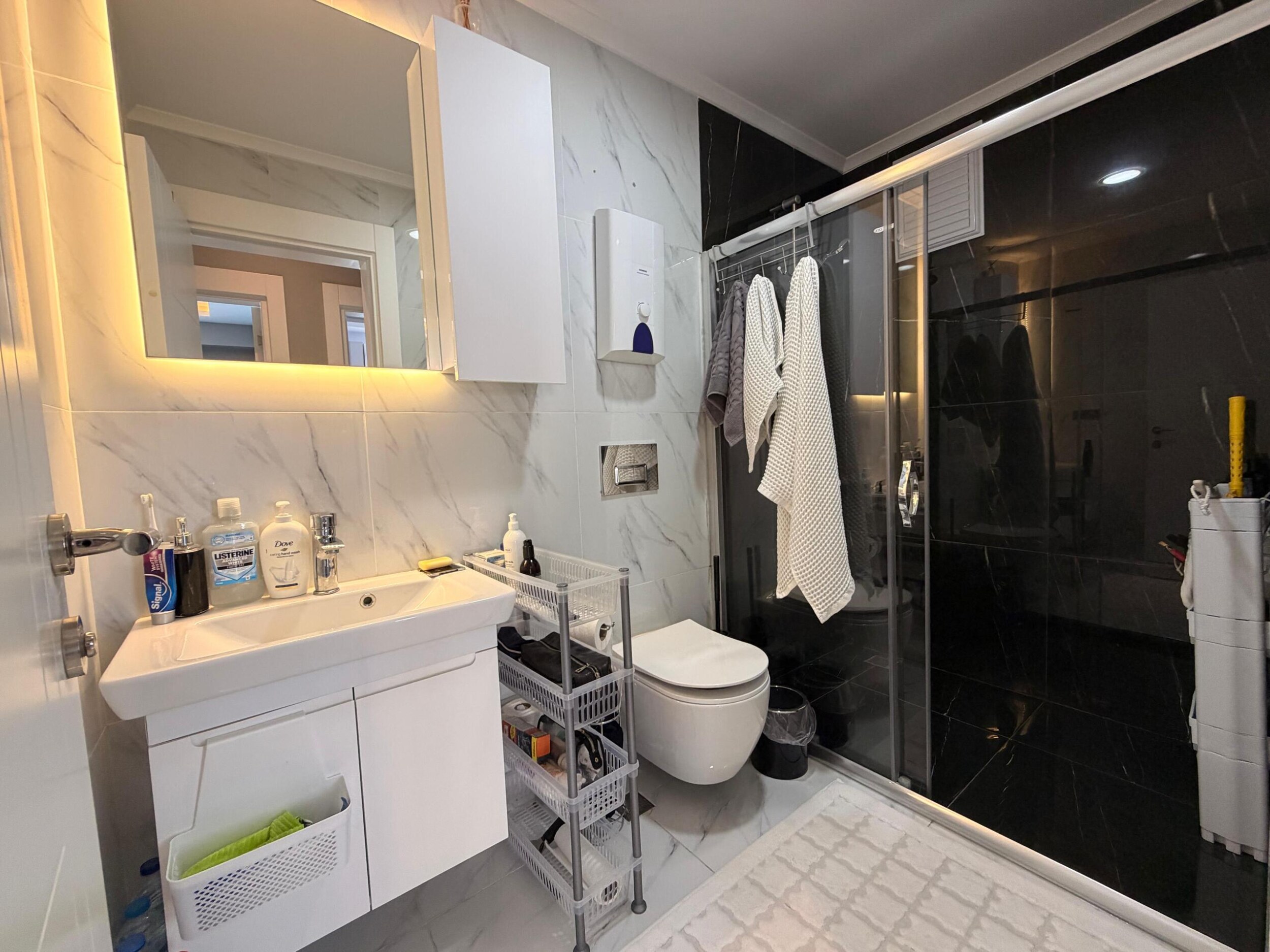 Apartman dairesinin banyosunda cam duş kabini, siyah fayans ve ışıklı aynalı dolap