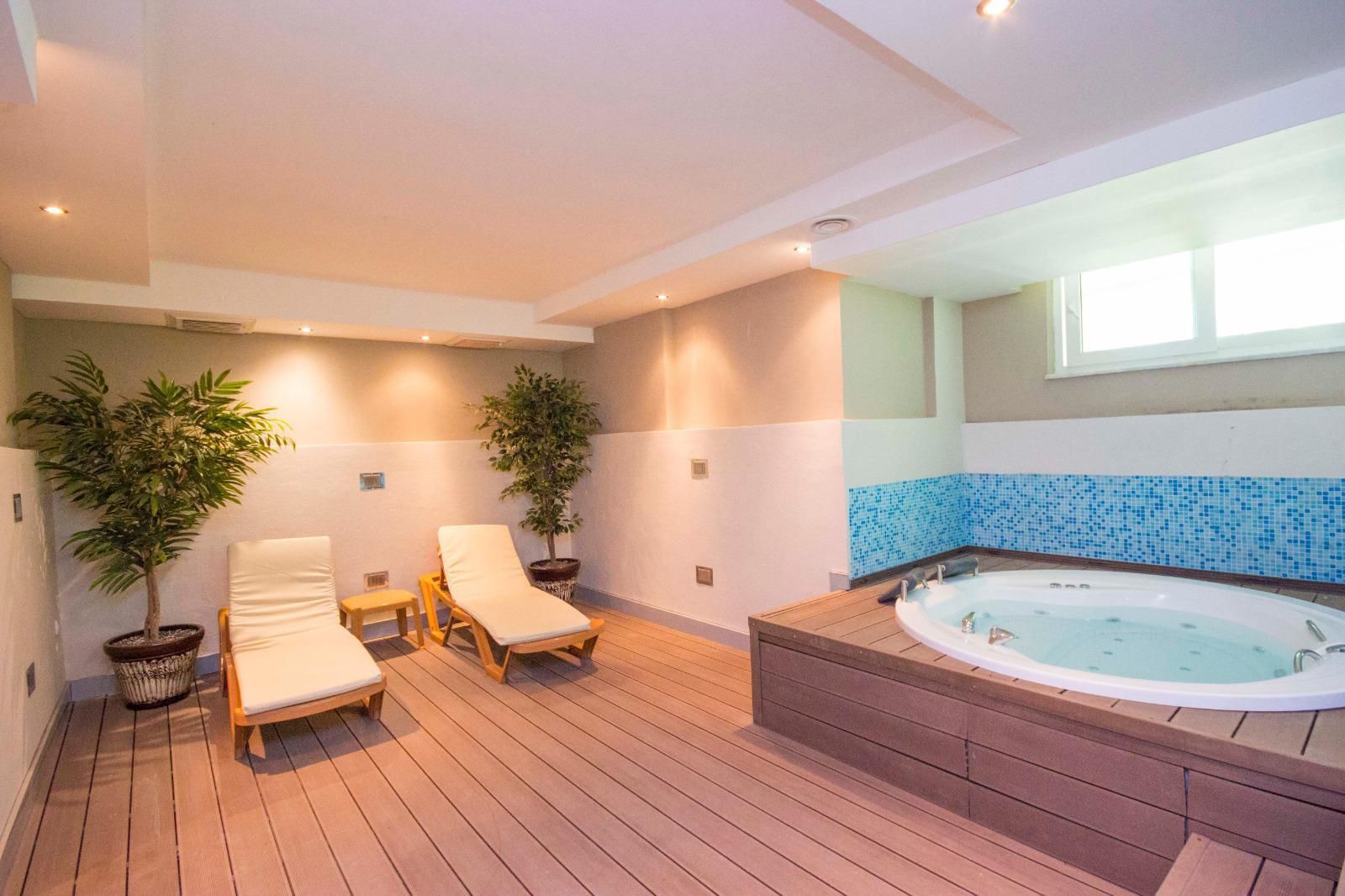 Spa alanında ahşap platformlu yuvarlak jakuzi, iki beyaz şezlong ve mavi mozaik kaplama
