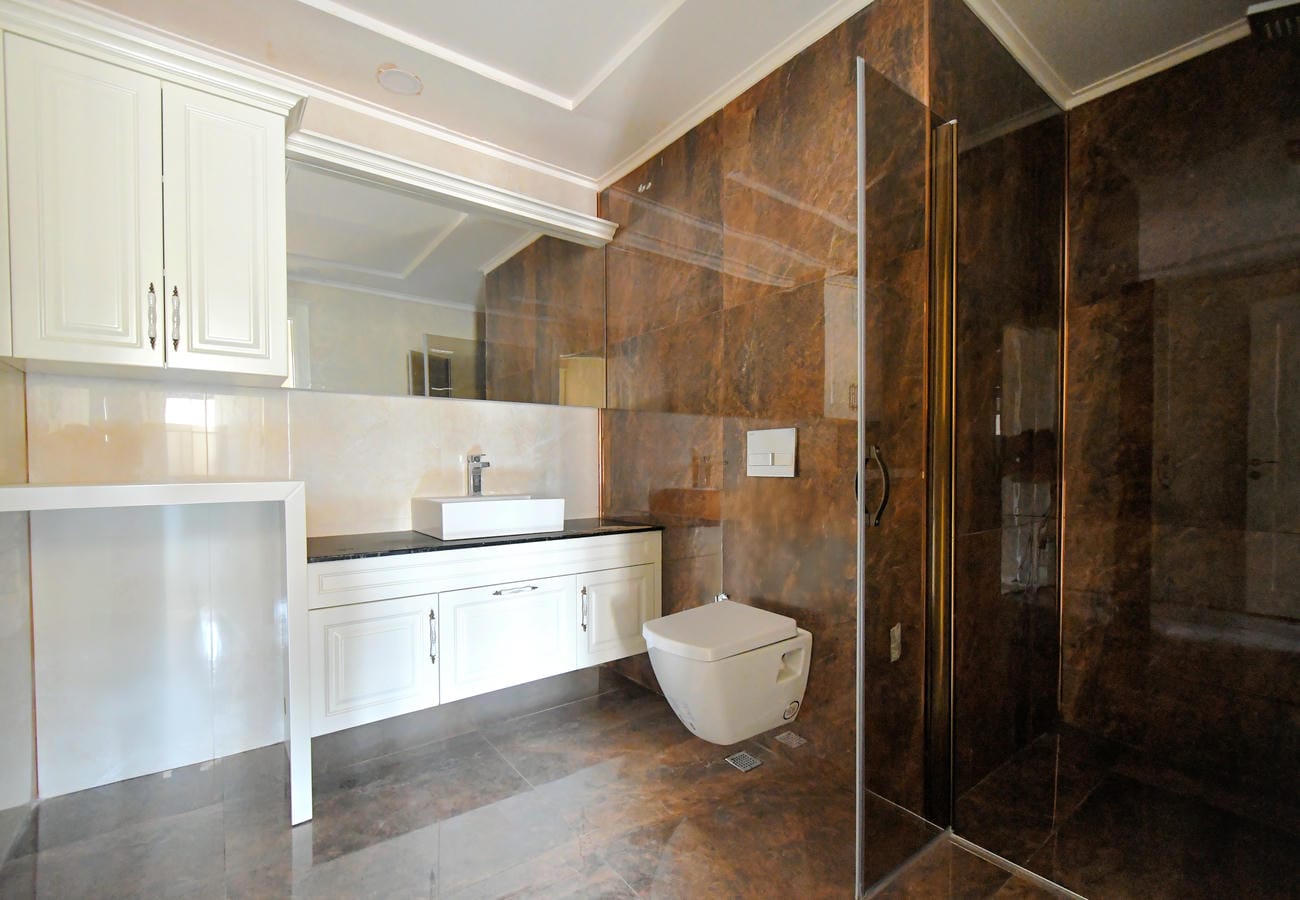 Alanya’da penthouse banyosunda kahverengi mermer duvarlar, cam duş bölmesi ve beyaz dolap