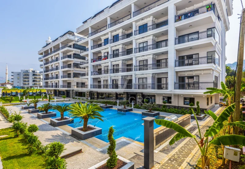 Beyaz konut blokları önündeki palmiyeli açık havuz: Kargıcak’ta satılık 3+1 penthouse, Alanya, Türkiye — New Level Group