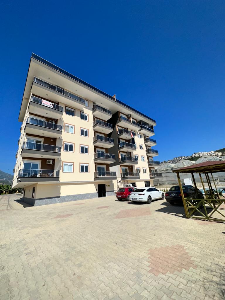 Cam korkuluklu beş katlı bina önünde açık otopark, Alanya’da dağlar eşliğinde