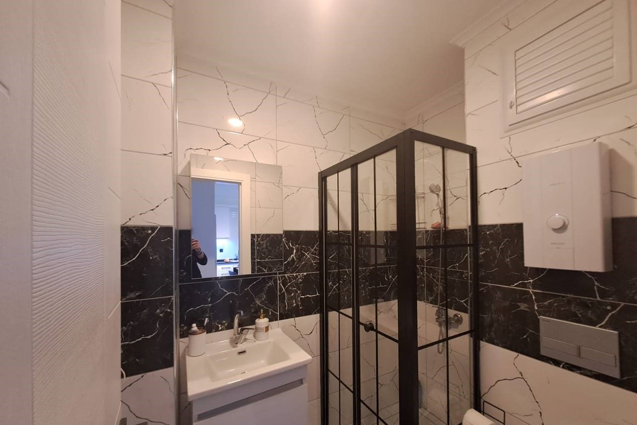 Cam duş kabinli banyo, dairede beyaz lavabo dolabı ve mermer desenli siyah beyaz fayanslarla