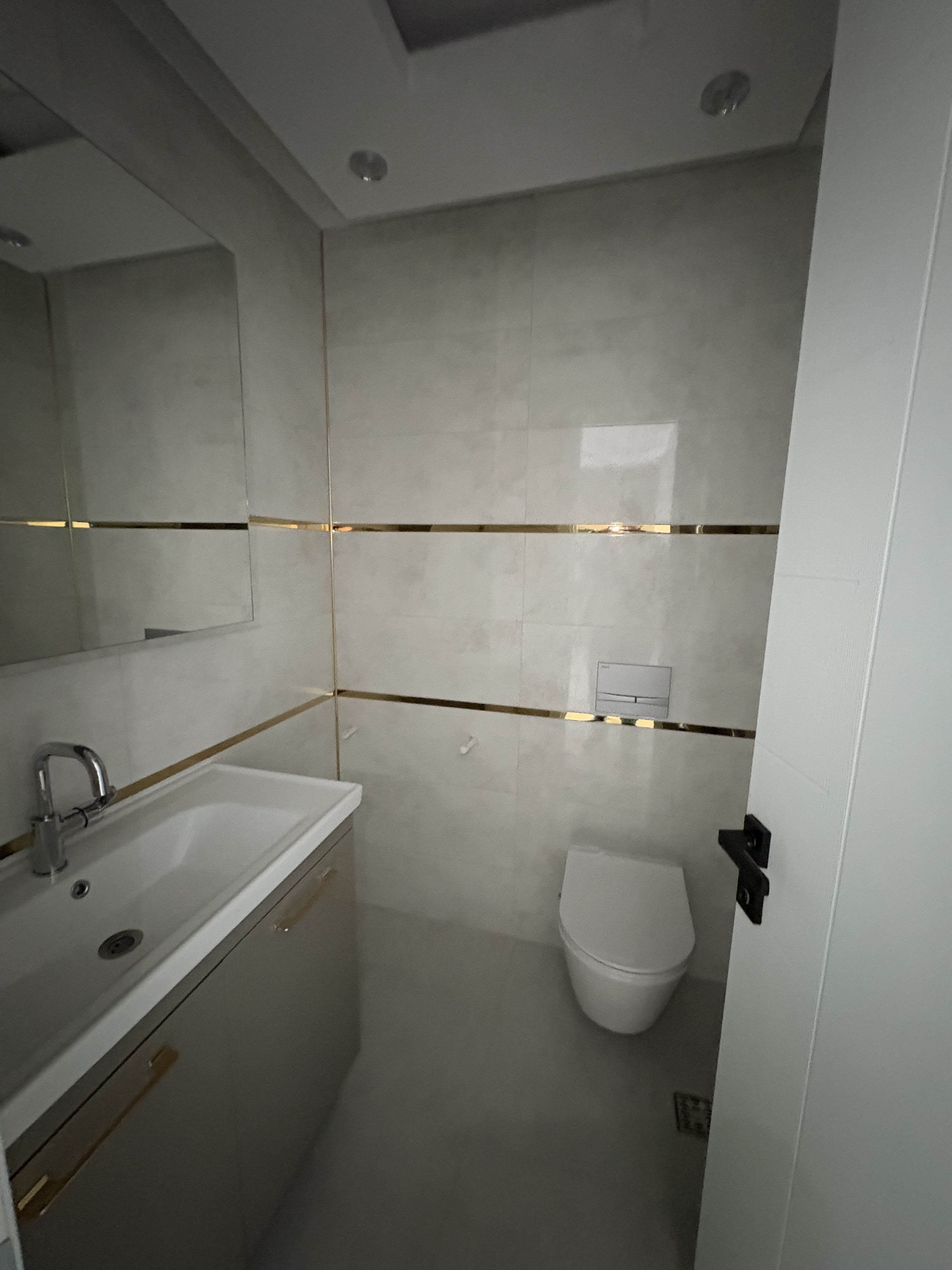 Aynalı duvarlı banyoda dolaplı lavabo, asma klozet ve altın renkli şeritler