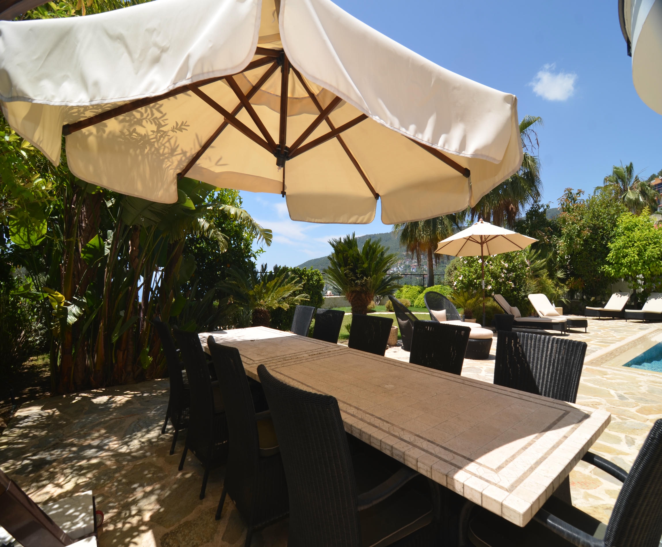 Long outdoor dining table with wicker chairs and an umbrella beside the pool at a house for sale in Turkey