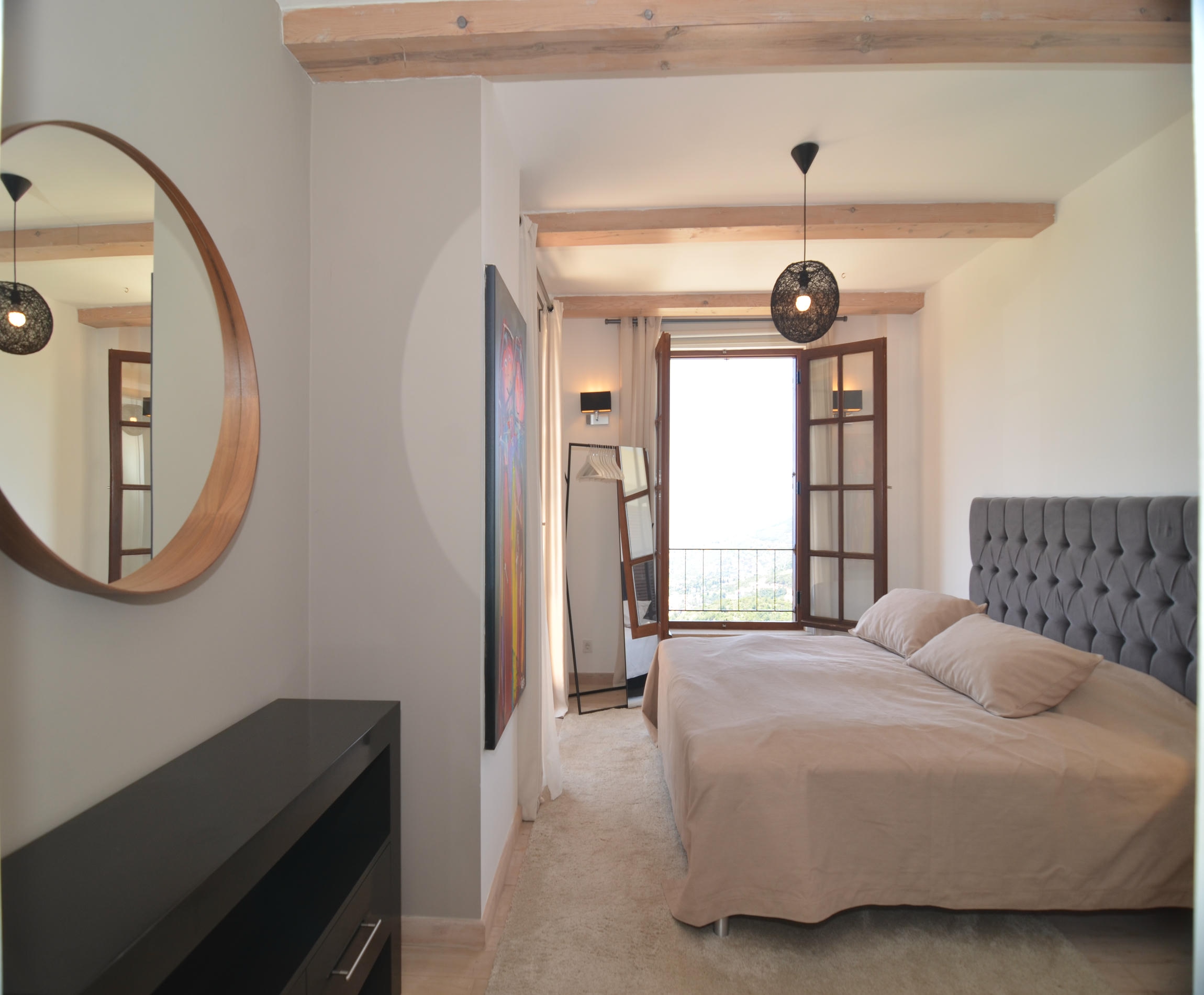 Bedroom with a gray quilted headboard, round mirror and timber beams in a villa