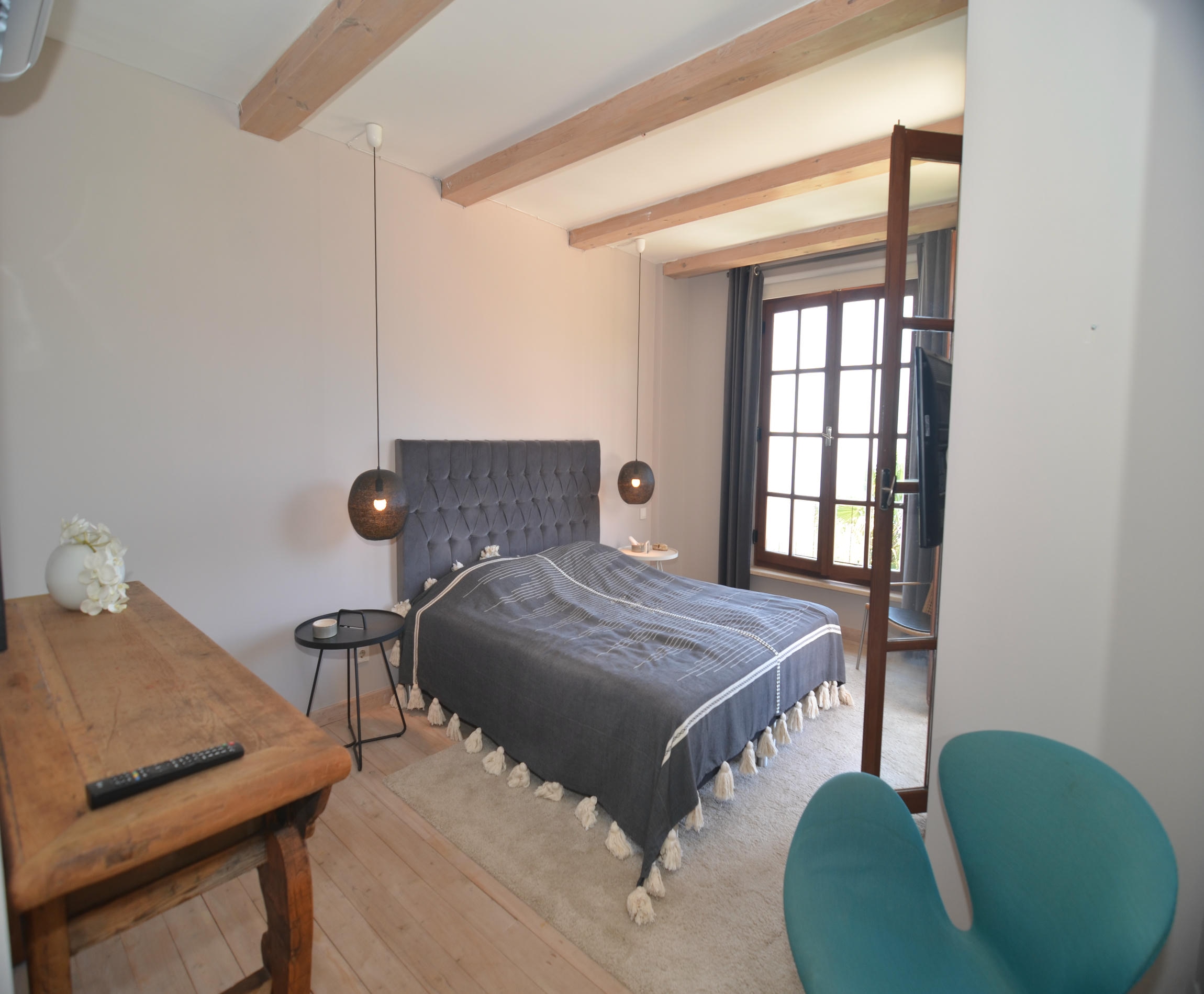 Bedroom with a gray quilted headboard, timber beams and turquoise armchair in a villa