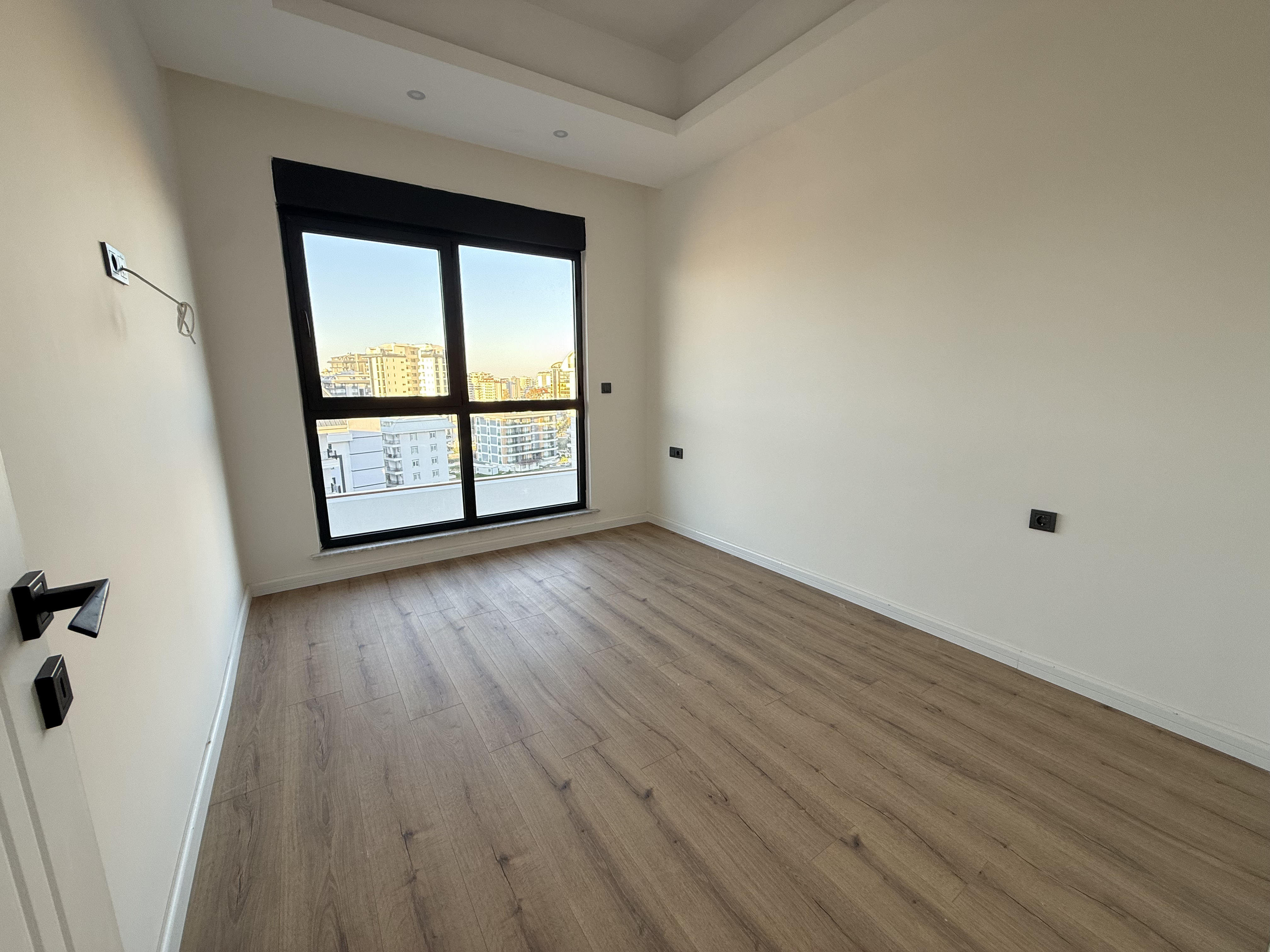 Empty bedroom with a wide window and wood-effect laminate overlooking city buildings