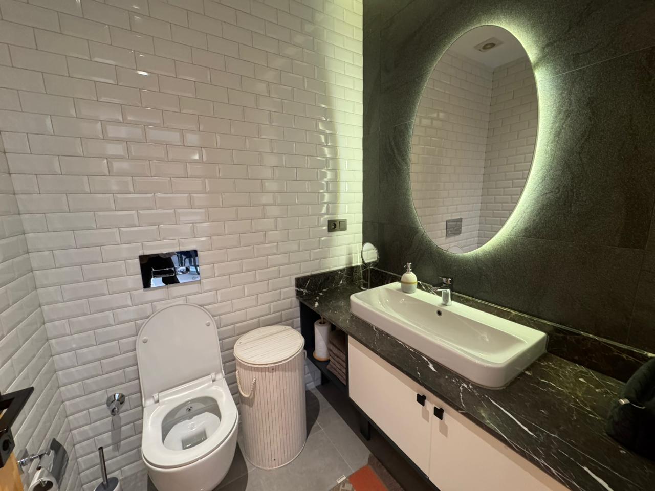 Beyaz metro fayanslı banyo ve ışıklı yuvarlak ayna Alanya’daki daireli konutta