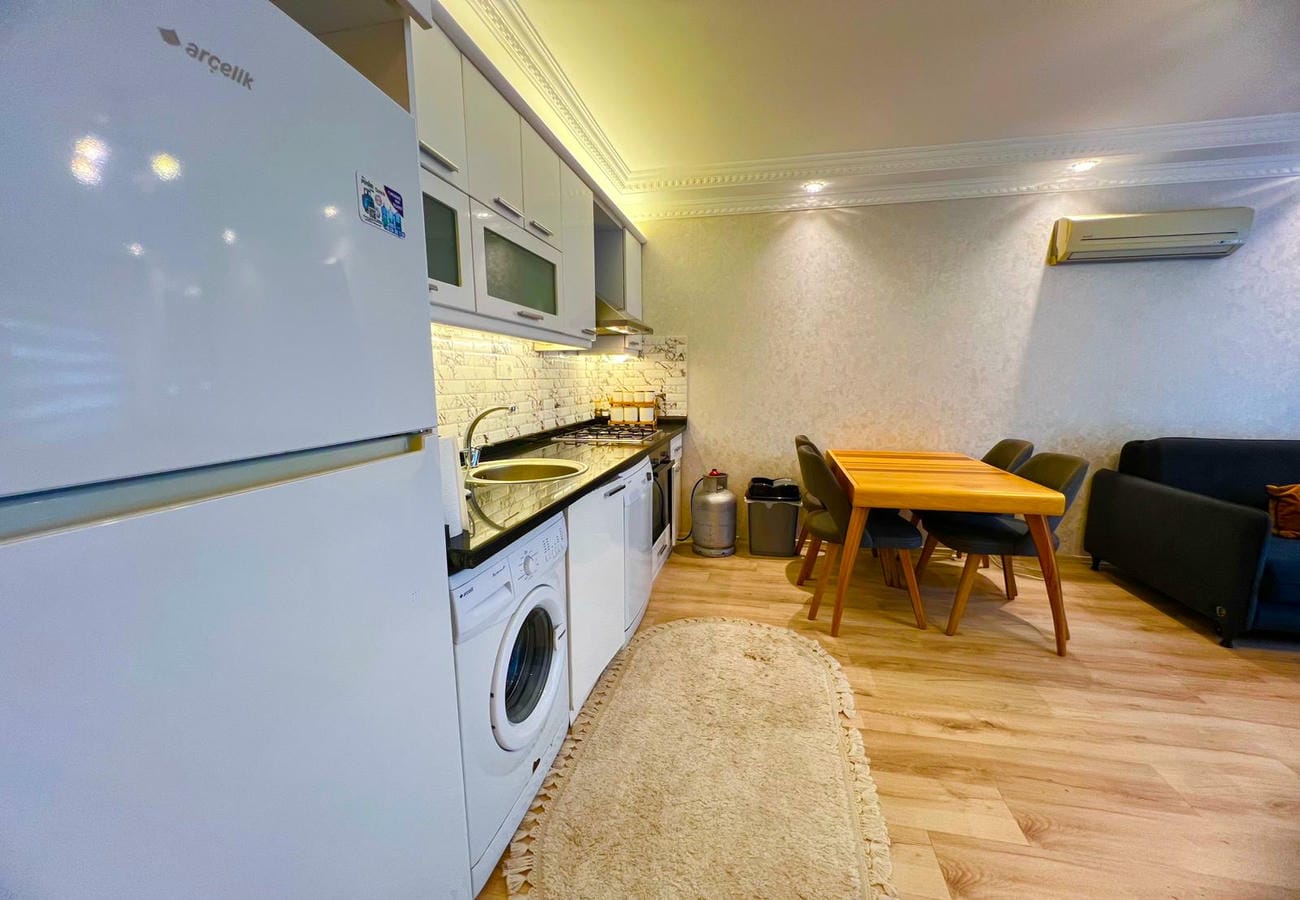Beyaz buzdolaplı mutfak-salon, ahşap yemek masası ve siyah kanepeli konutta
