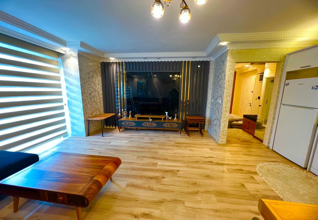 Ahşap sehpalı mutfak-salon, mavi köşe kanepe ve panjurlu panoramik pencereyle konutta