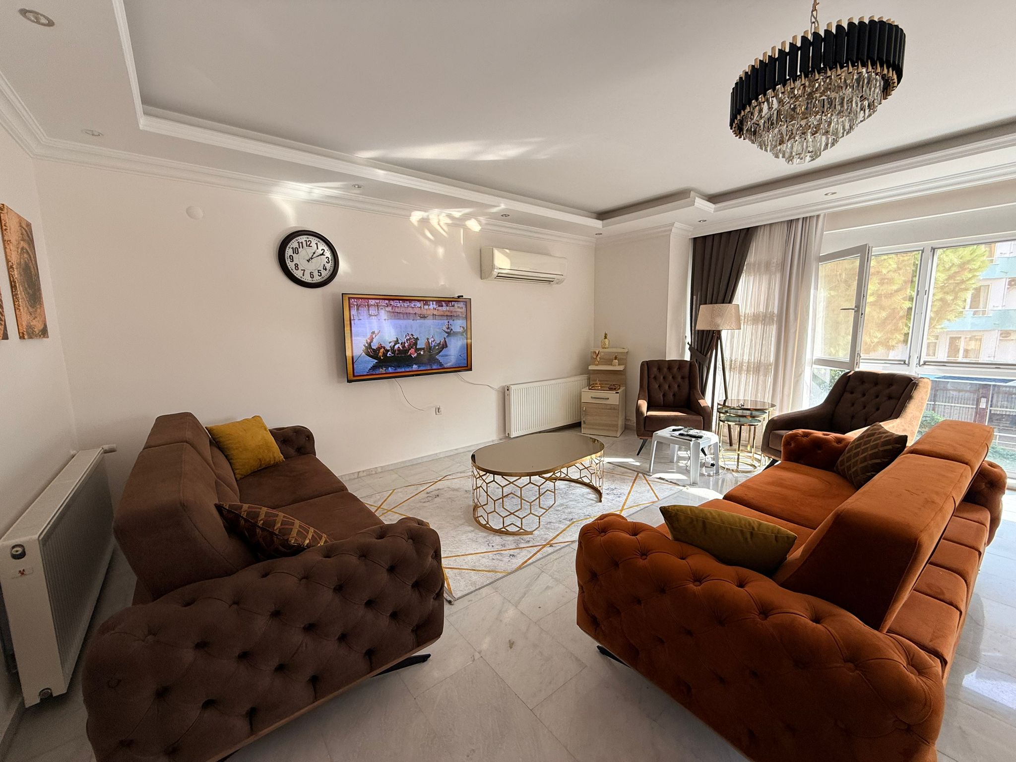 Turuncu ve kahverengi koltuklu salon, satışta olan dairede televizyon ve kristal avizeyle düzenlenmiş