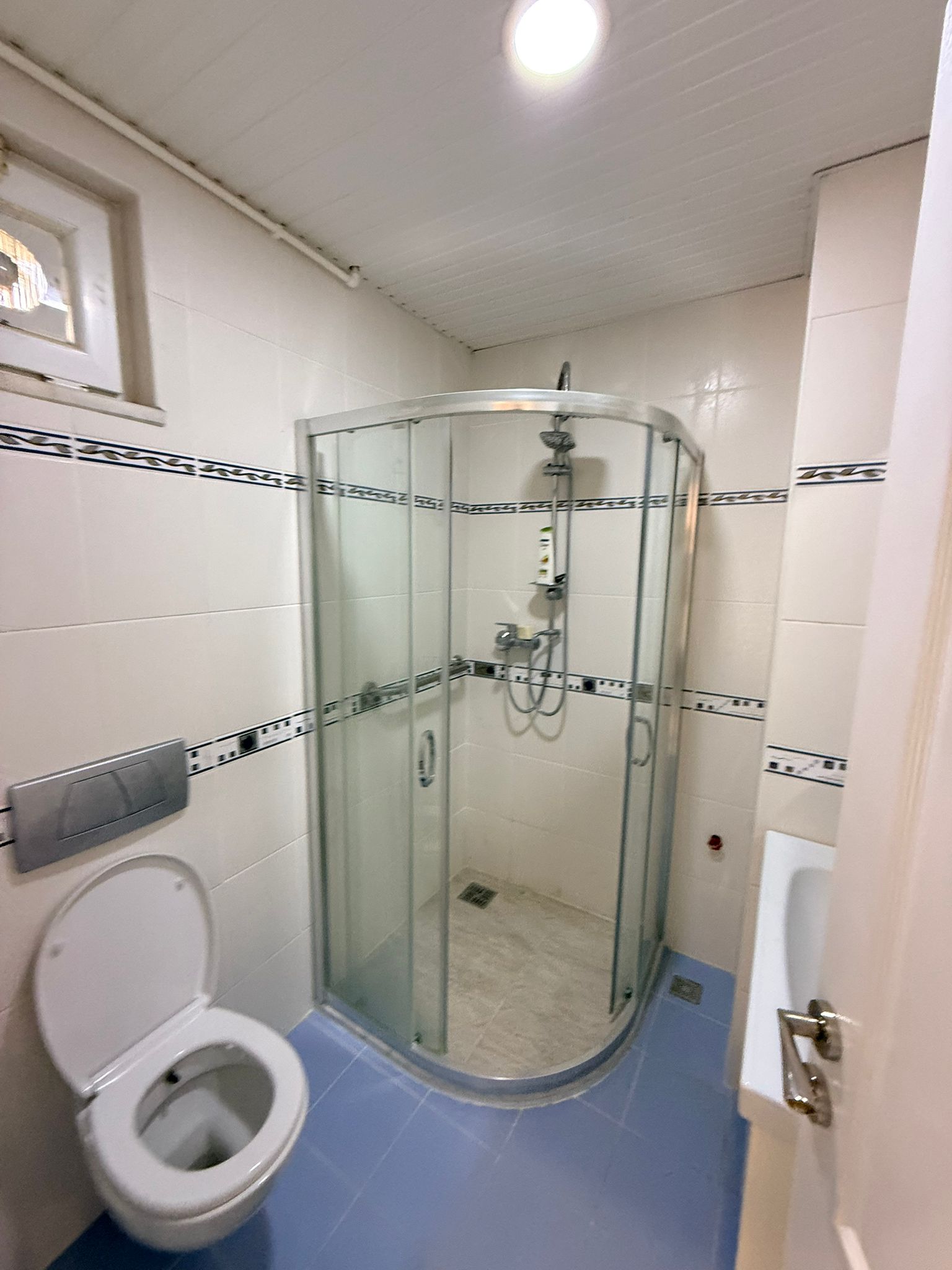 Yuvarlak cam duş kabinli, klozetli ve mavi zemin fayanslı banyo dairede yer alıyor