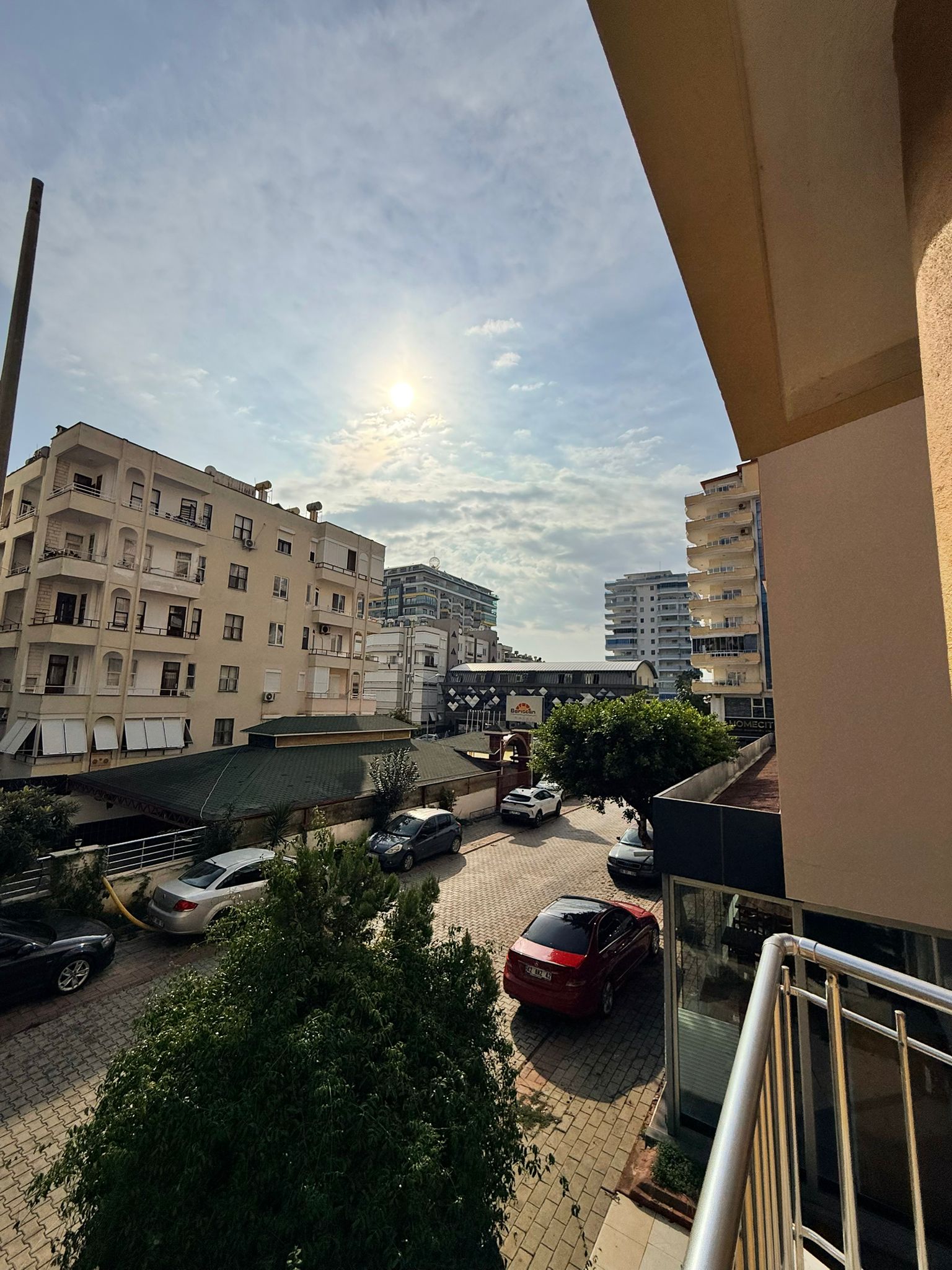 Terasın önünden şehir sokağı, konut binaları, ağaçlar ve park etmiş araçlarla görünüyor