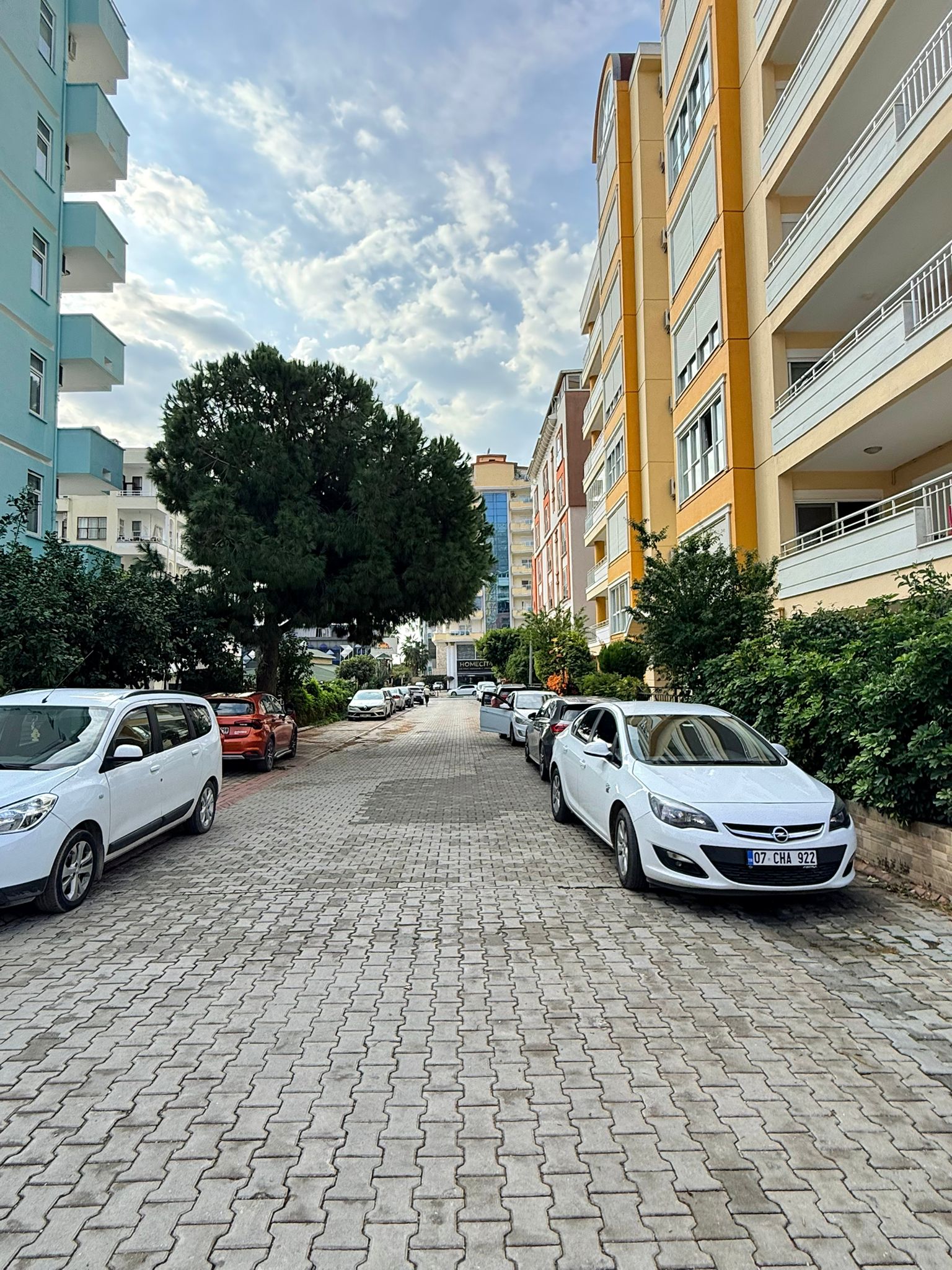 Mahmutlar'da satılık konutun yanında çalılarla çevrili taş döşeli iç yol ve park etmiş araçlar
