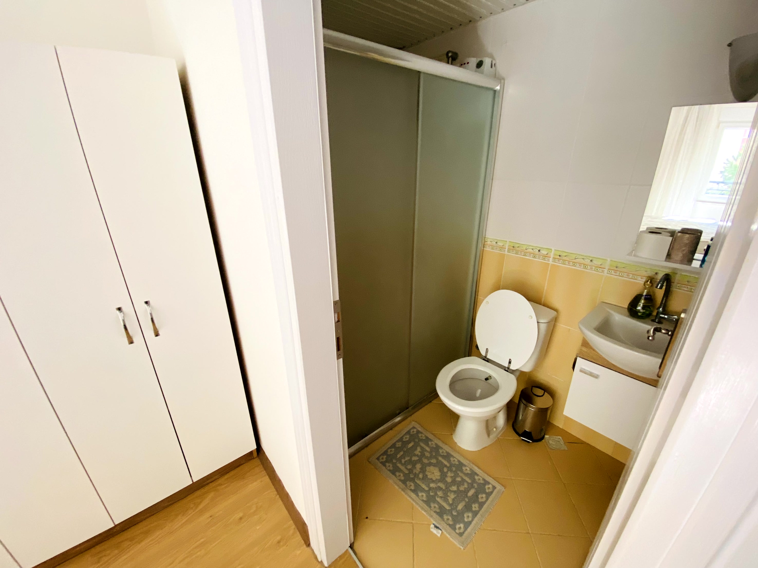 Cam kapılı duş kabinli banyo Mahmutlar’daki dairede aynalı lavabo ve sarı-beyaz fayanslarla