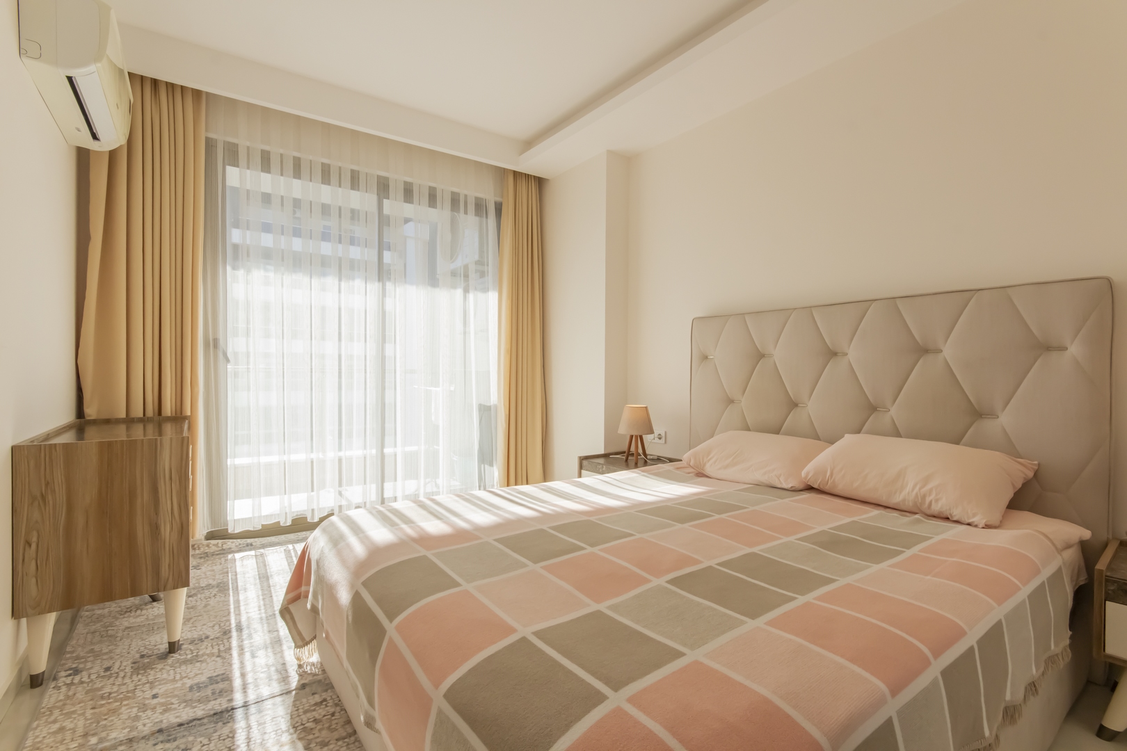 Konutun yatak odasında yüksek kapitone başlık, panoramik pencere, sarı perdeler ve ahşap şifonyer
