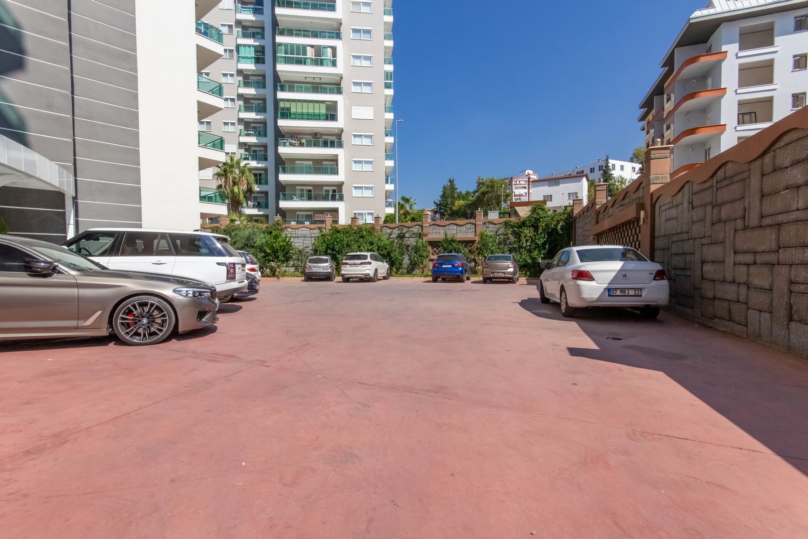 Mahmutlar’daki dairelerin yanında kırmızı zeminli açık otopark, park edilmiş araçlar ve taş çit
