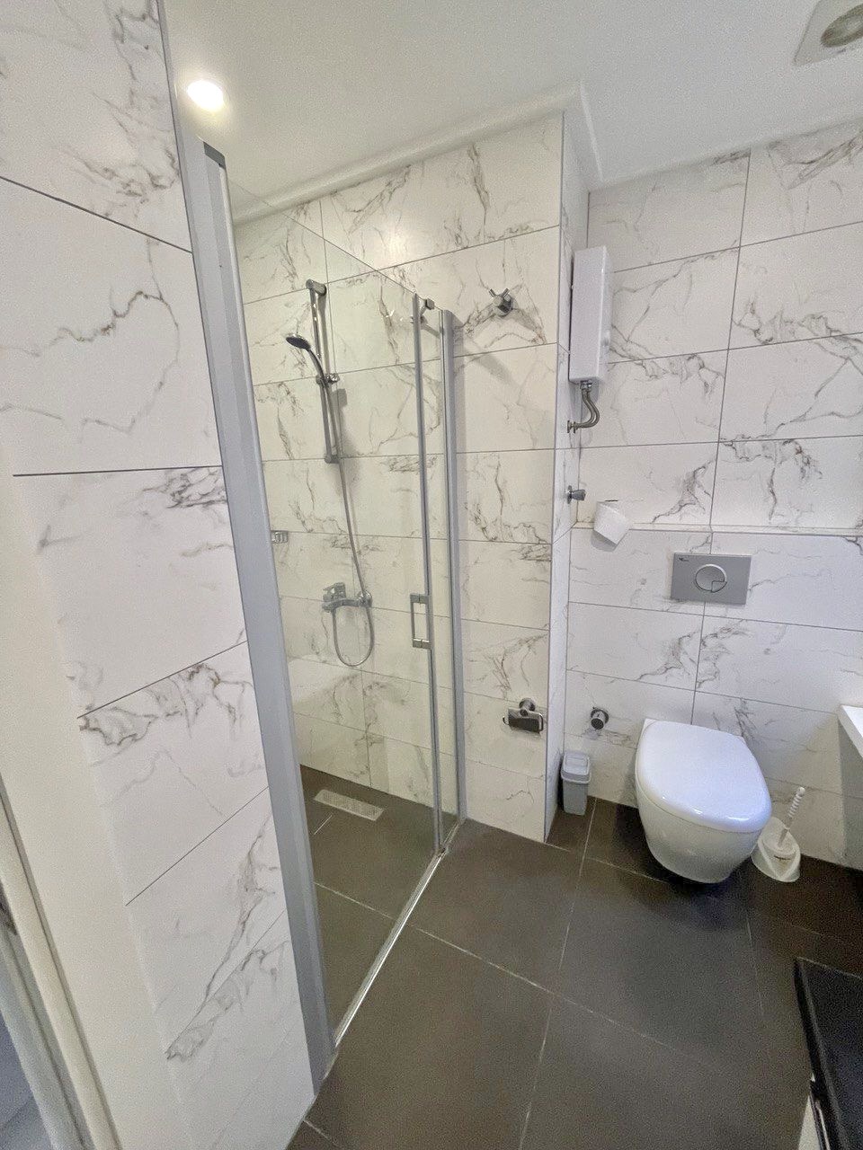 Cam duş kabinli banyo, dairenin mermer kaplı duvarları ve asma klozetiyle