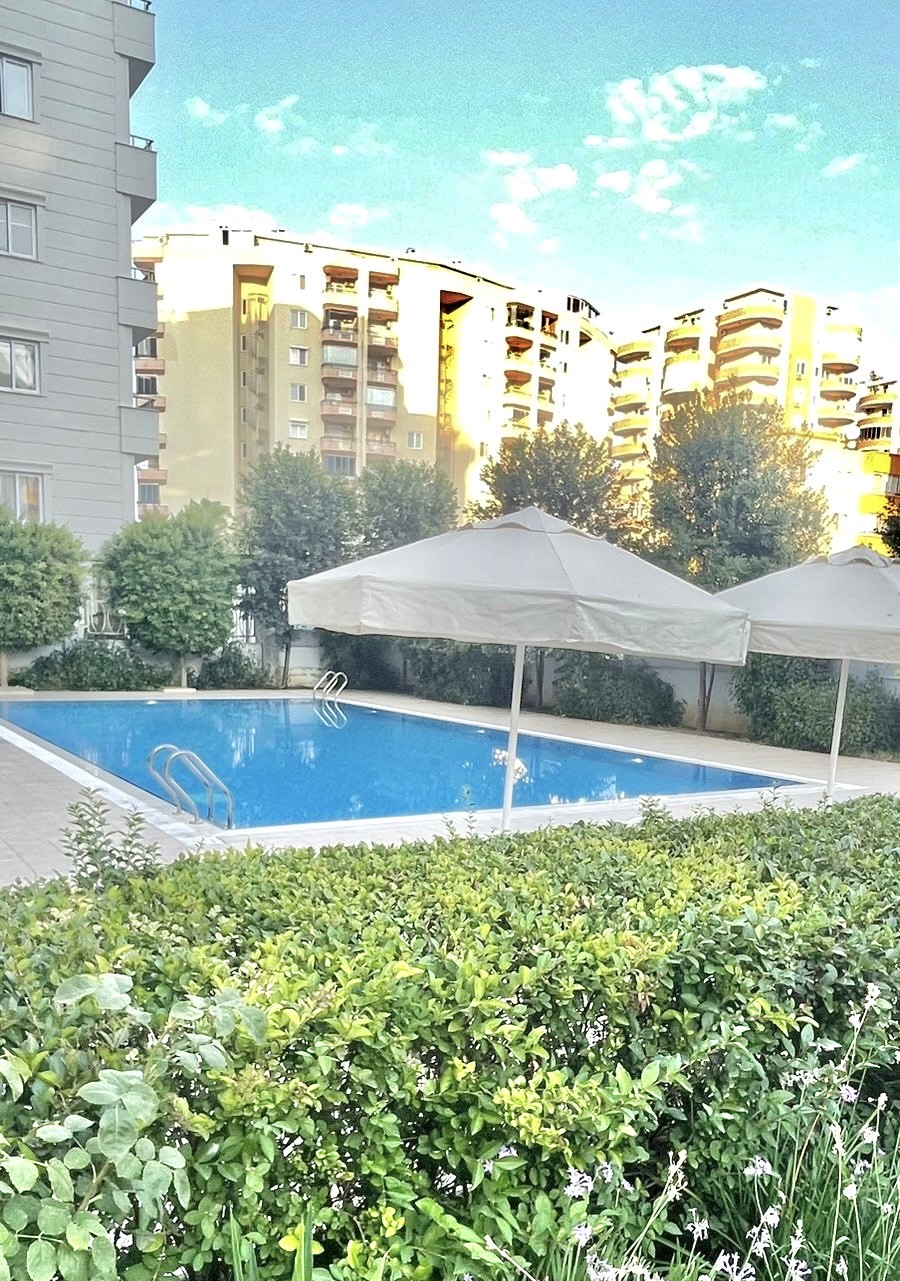 Beyaz şemsiyeli açık havuz ve yeşil çit, satılık dairelerin bulunduğu rezidansta