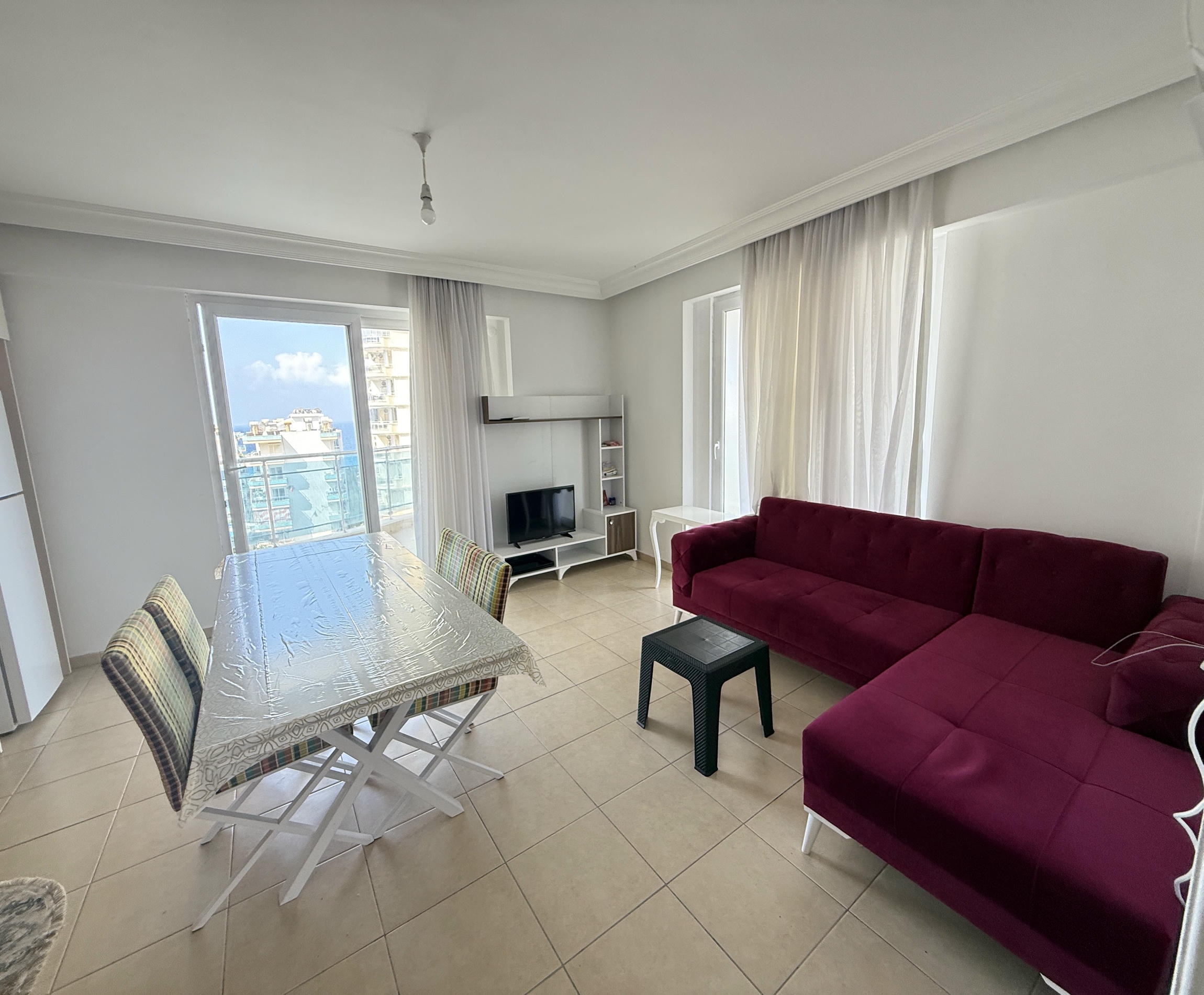 Bordo köşe koltuklu mutfak-salon satılık dairede yüksek pencereler ve cam balkon görülüyor