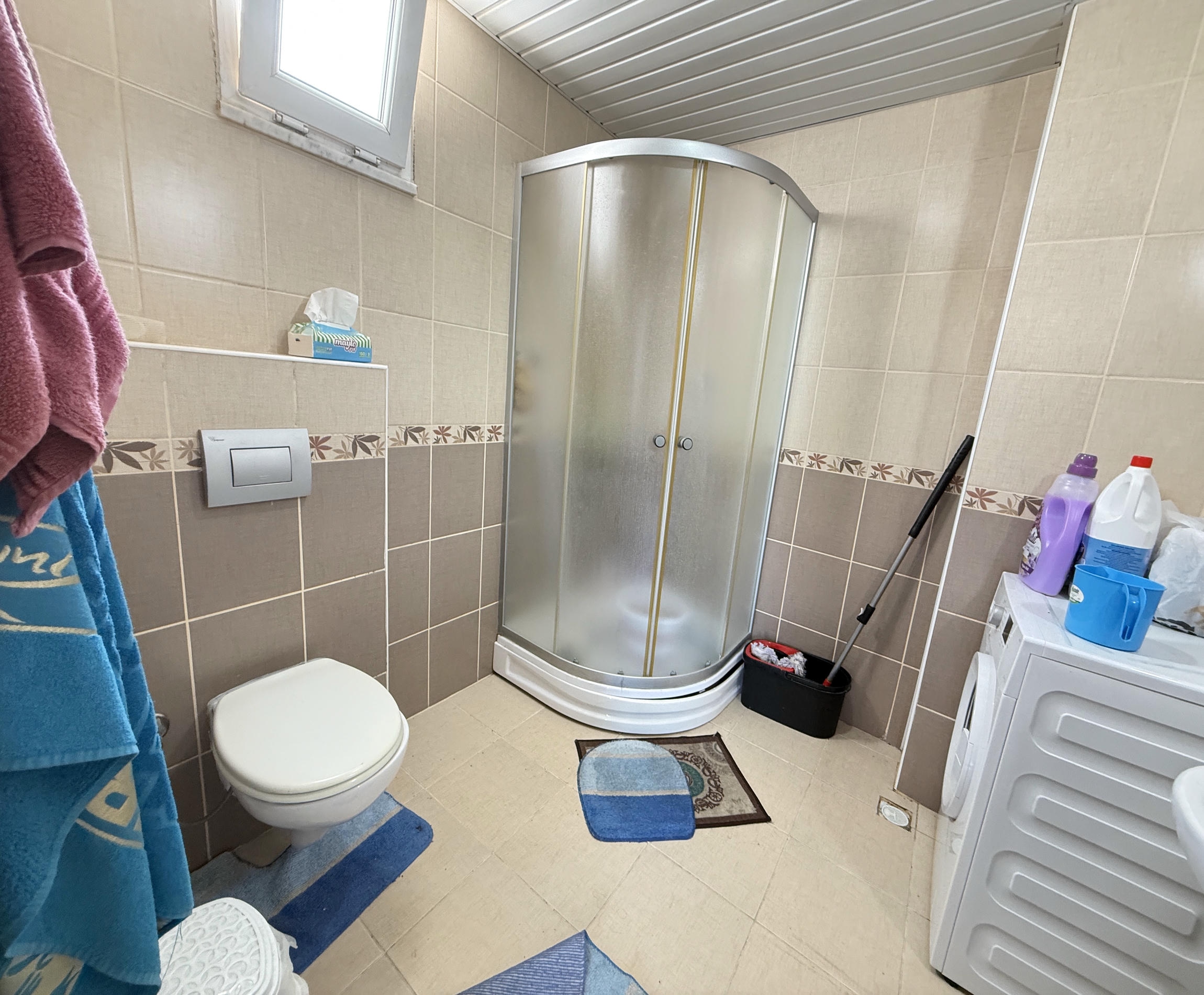 Duşakabinli banyo Mahmutlar’daki dairede çamaşır makinesi ve bej fayanslarla düzenlenmiş