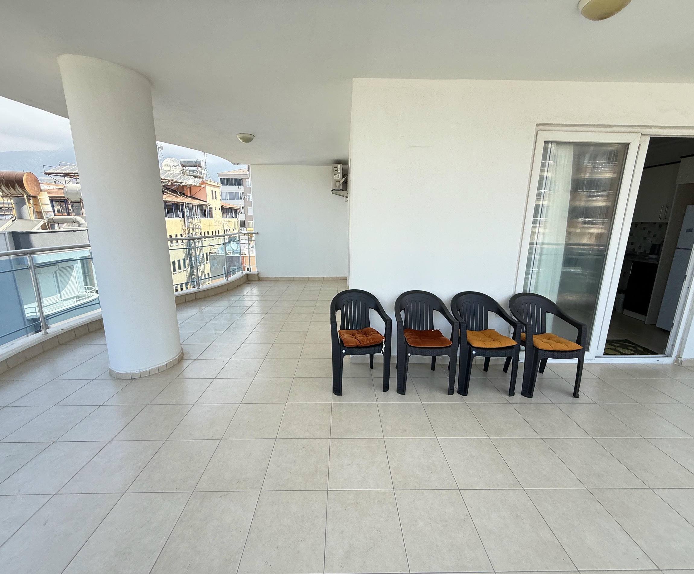 Geniş kapalı teras, apartmanda siyah sandalyeler ve cam korkulukla şehir binalarına bakıyor