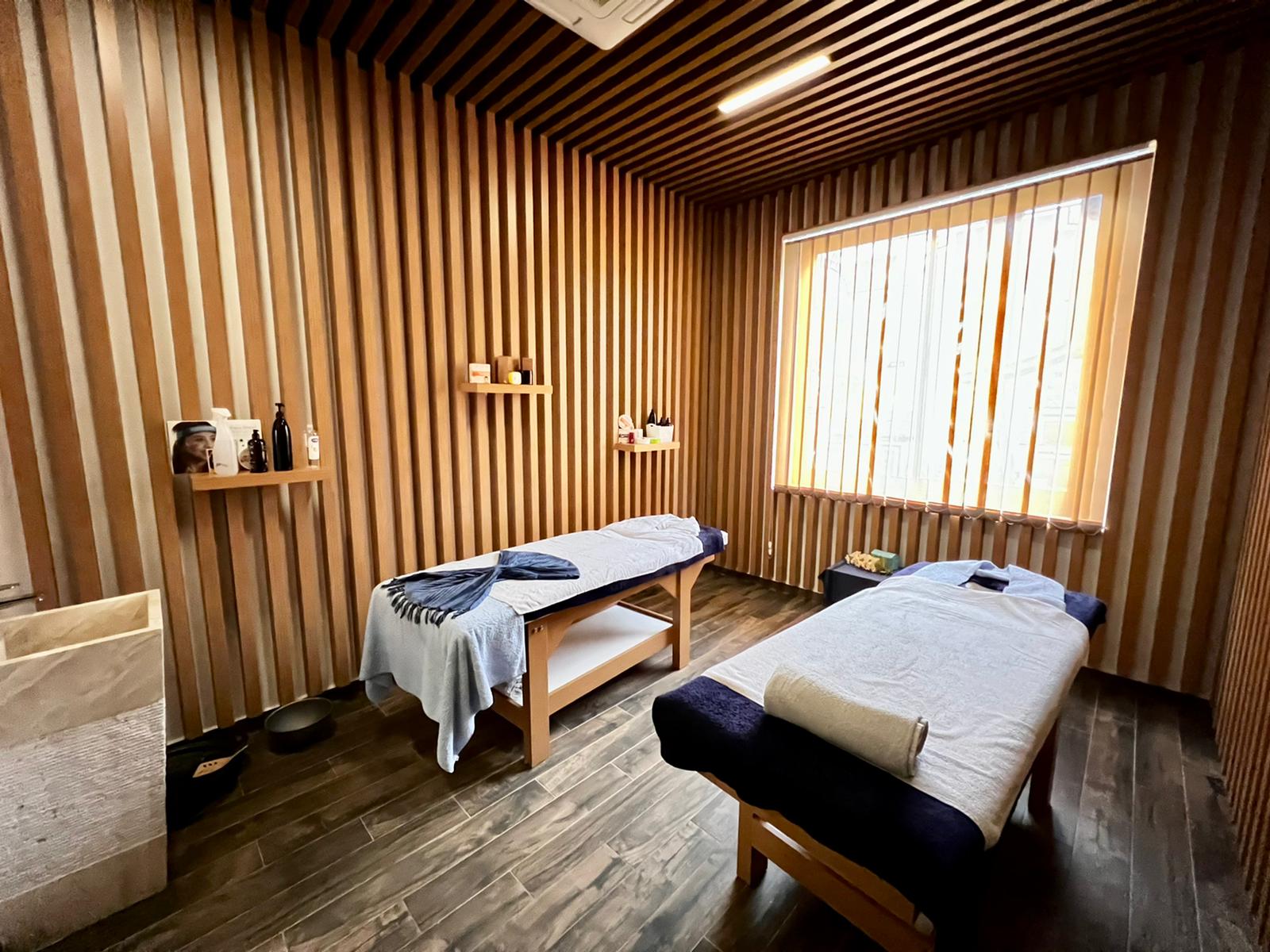 Spa area with two massage tables and wood slat walls beside a window with blinds