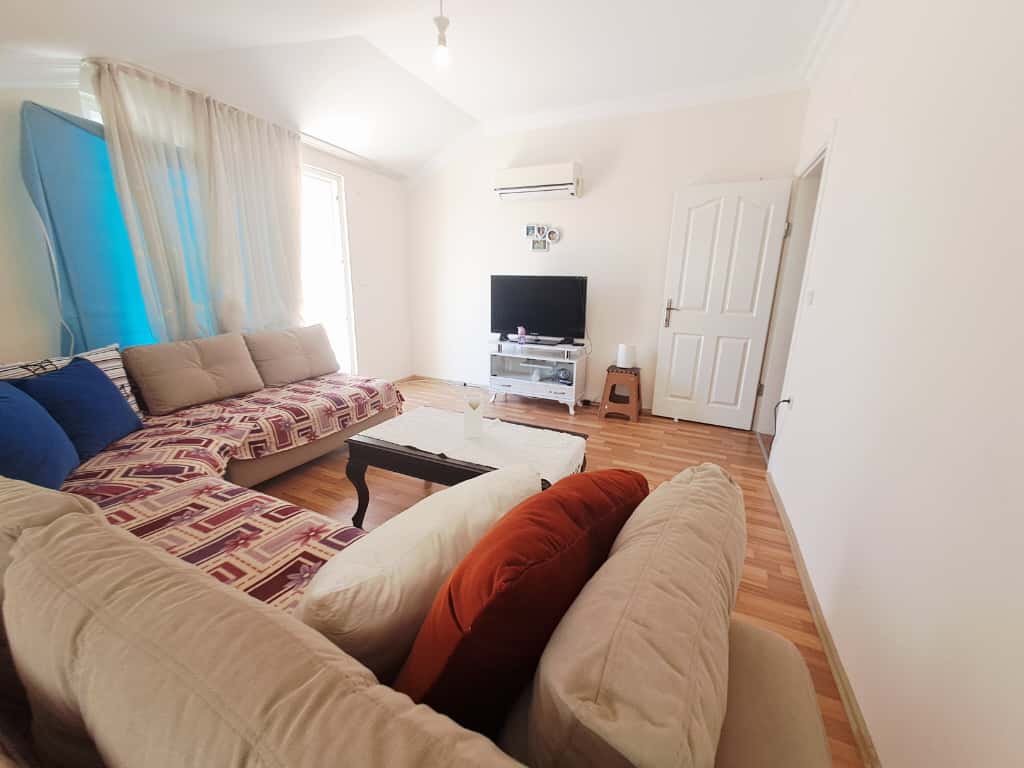 Alanya'da satılık evin salonunda geometrik örtülü köşe kanepe ve cam sehpada televizyon