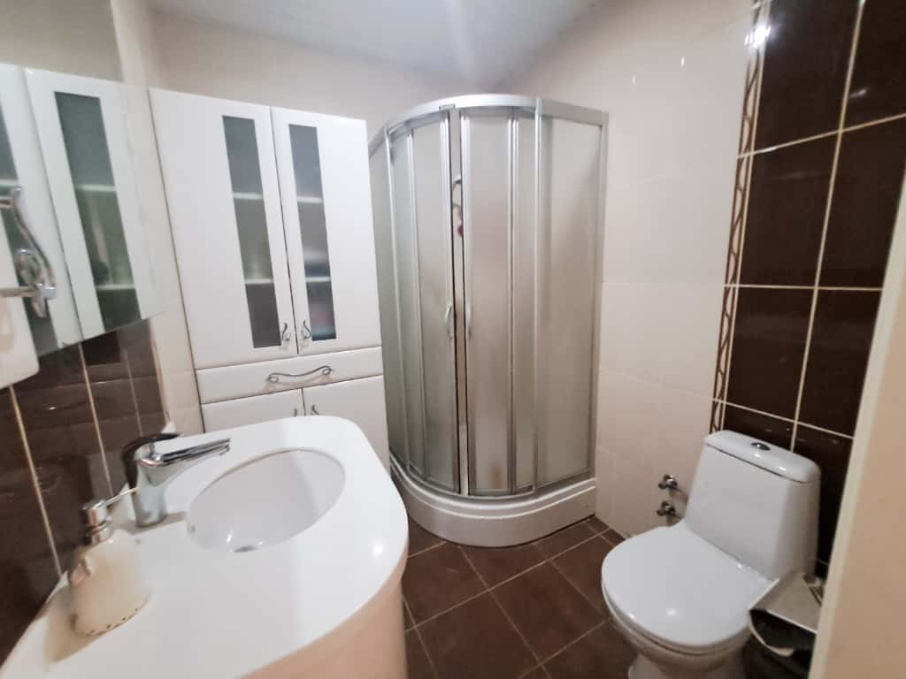 Çikcilli'deki banyoda mat kapılı köşe duş kabini, beyaz lavabo ve kahverengi fayanslar