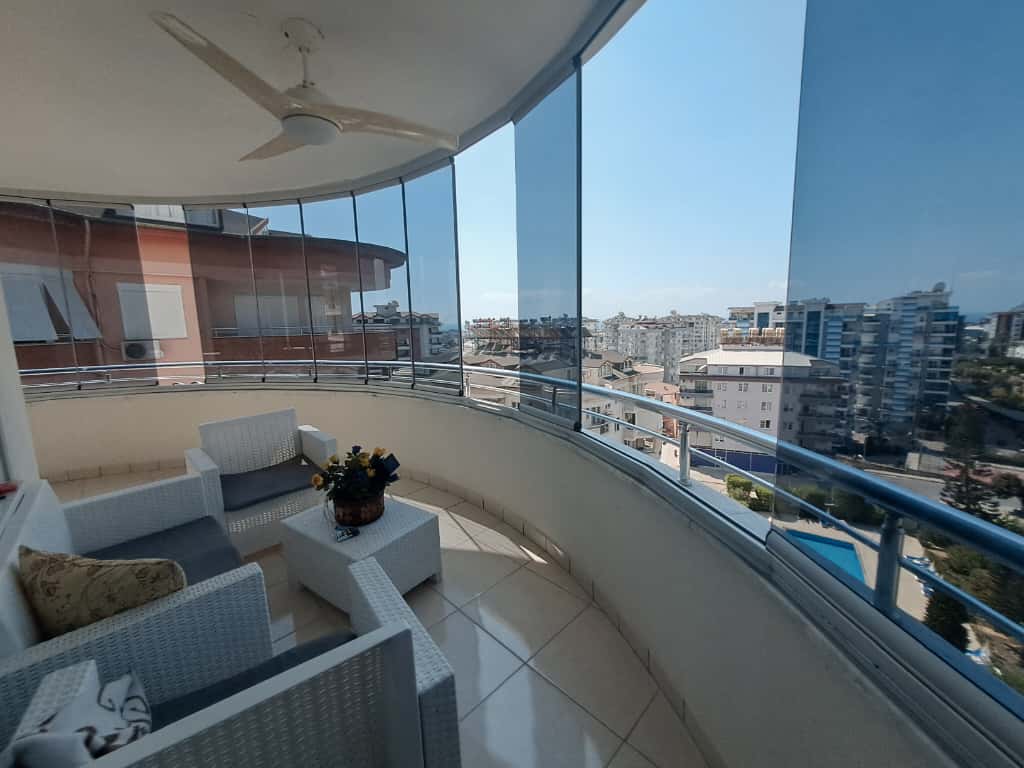 Alanya'daki 4+1 penthouse'un balkonunda beyaz hasır koltuklar, panoramik camlar ve tavan vantilatörü