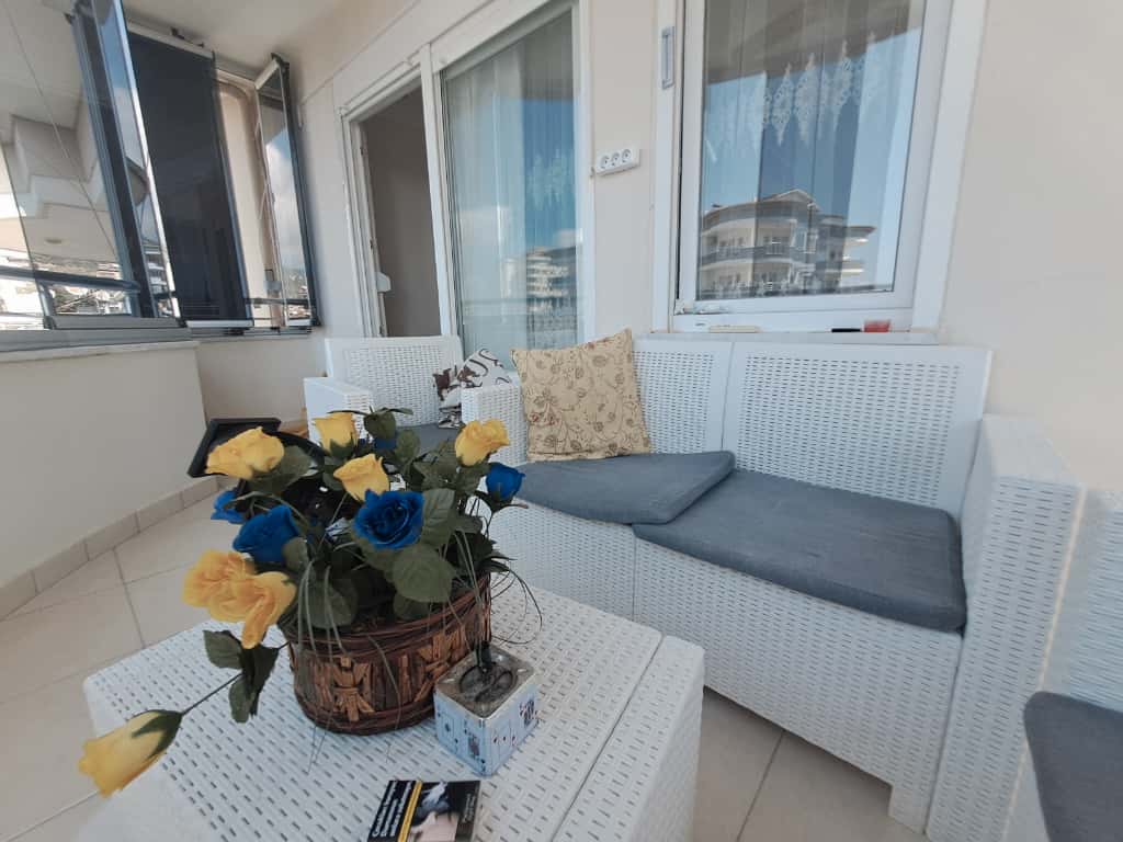Satılık evin balkonunda beyaz hasır kanepe, çiçek sepeti ve sürgülü cam kapılar