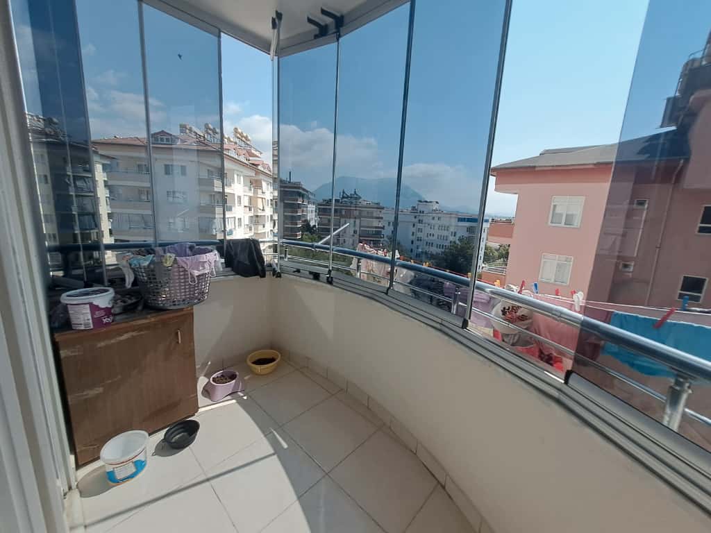 Alanya'daki camla kapalı köşe balkonda ahşap saklama dolabı, sepet ve komşu binalar manzarası