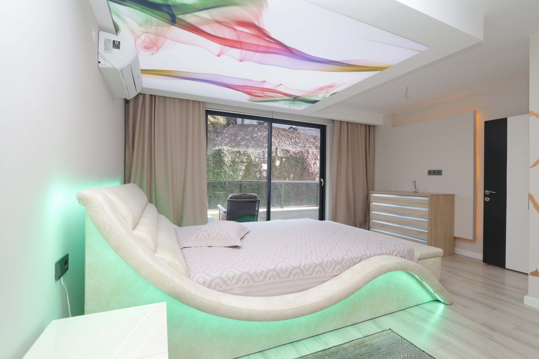 A bedroom with a curved upholstered headboard by a panoramic garden-facing window in a flat in Kargicak