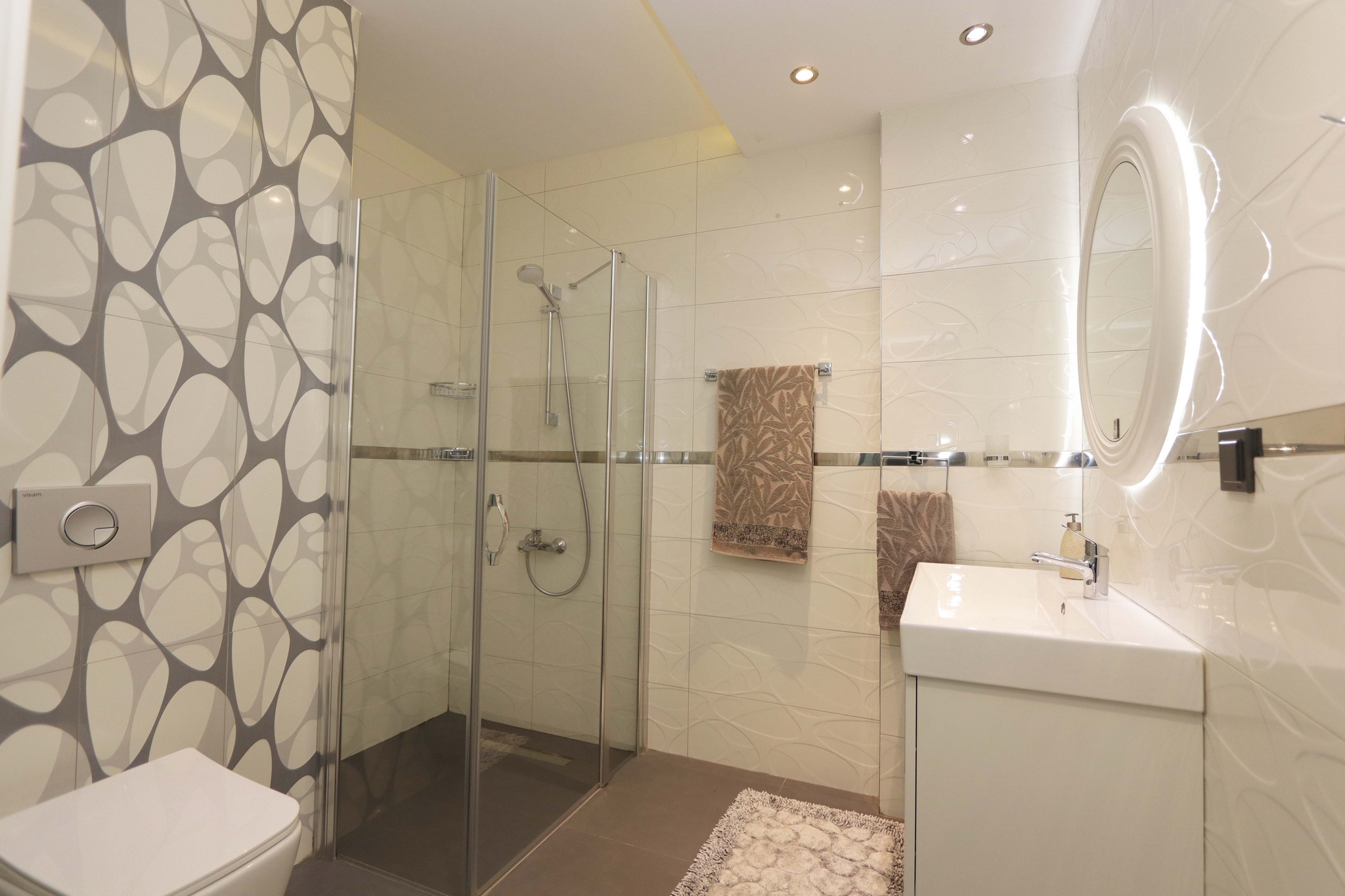 Bathroom with a glass shower enclosure, textured tile and round lit mirror in an apartment in Kargicak