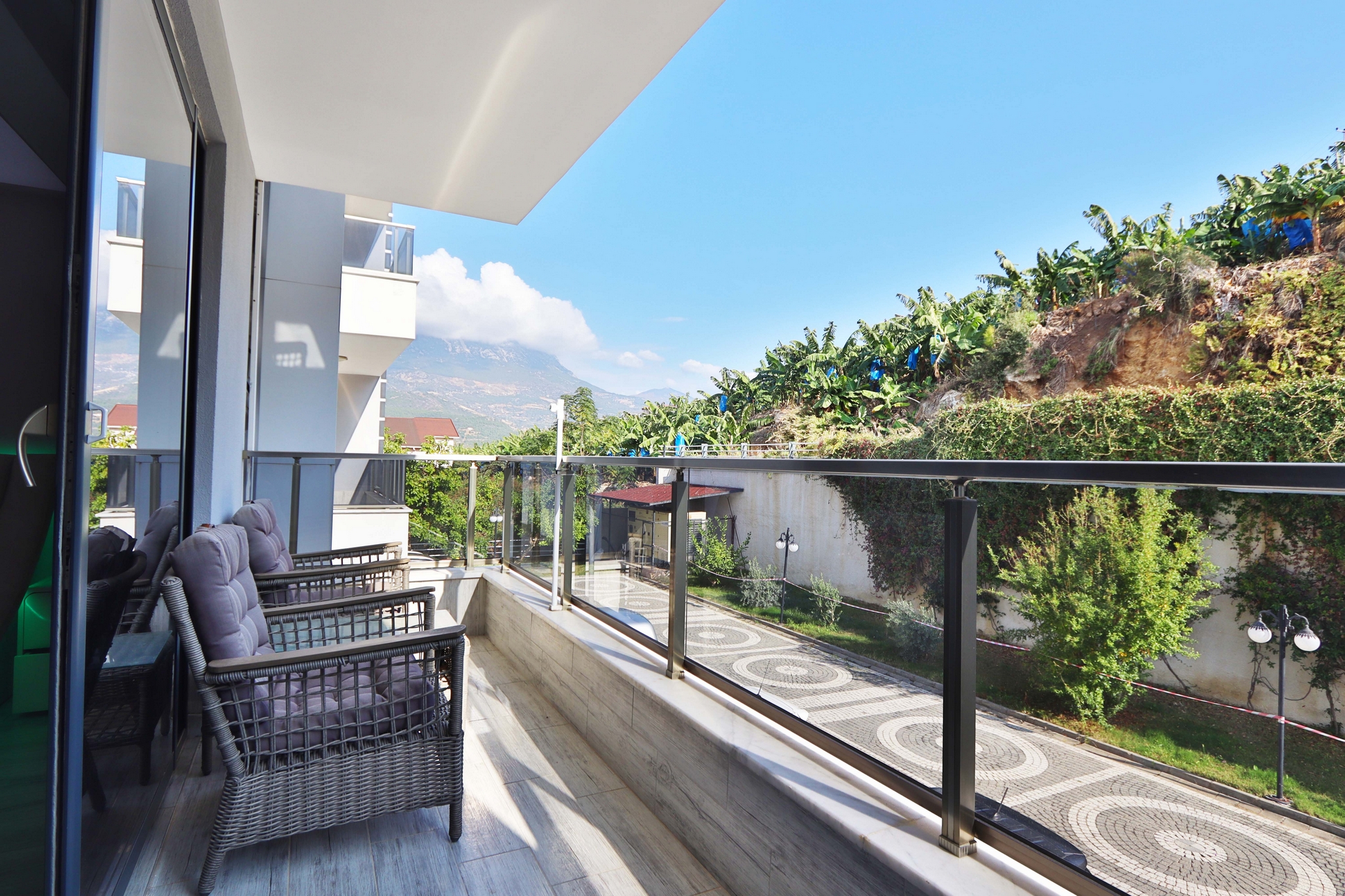 A balcony with glass railing, wicker chairs and banana plants on a slope beside a flat for sale in Turkey