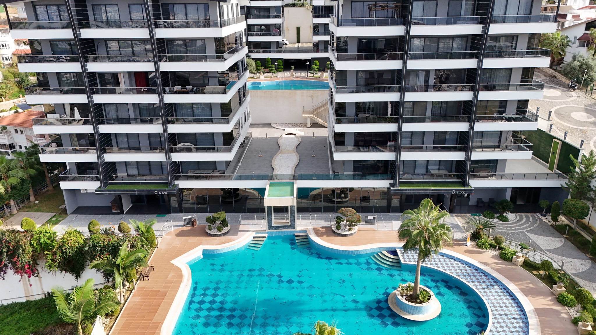 Open-air pool with a palm island and wooden decking beside balcony-fronted buildings where apartments are for sale in Turkey