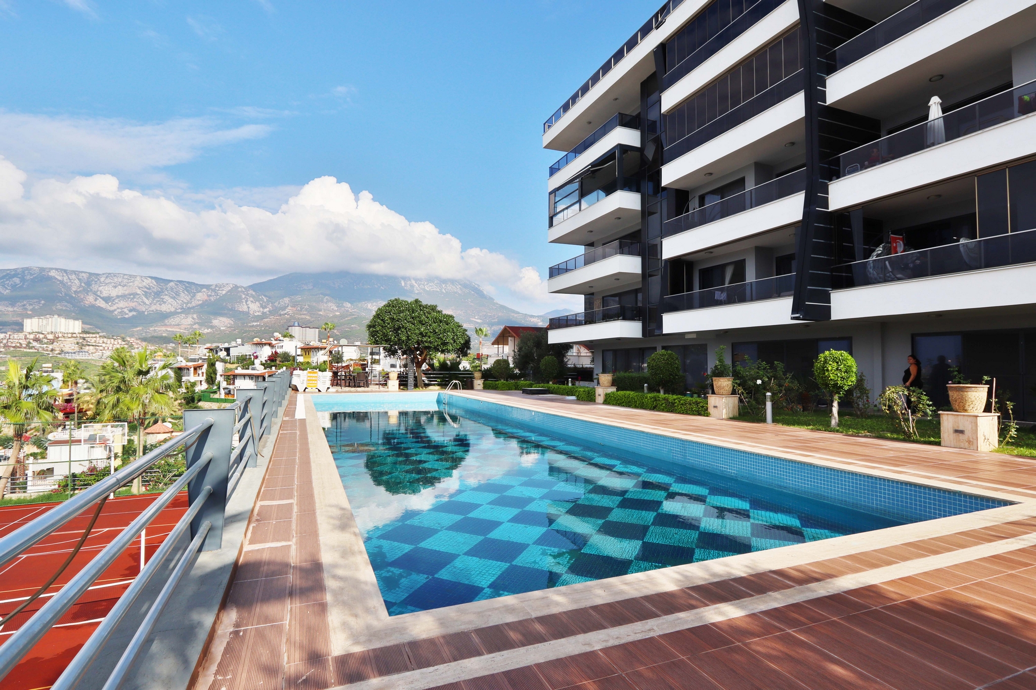 Rectangular outdoor pool with a wooden terrace and mountain backdrop in a complex with apartments in Kargicak
