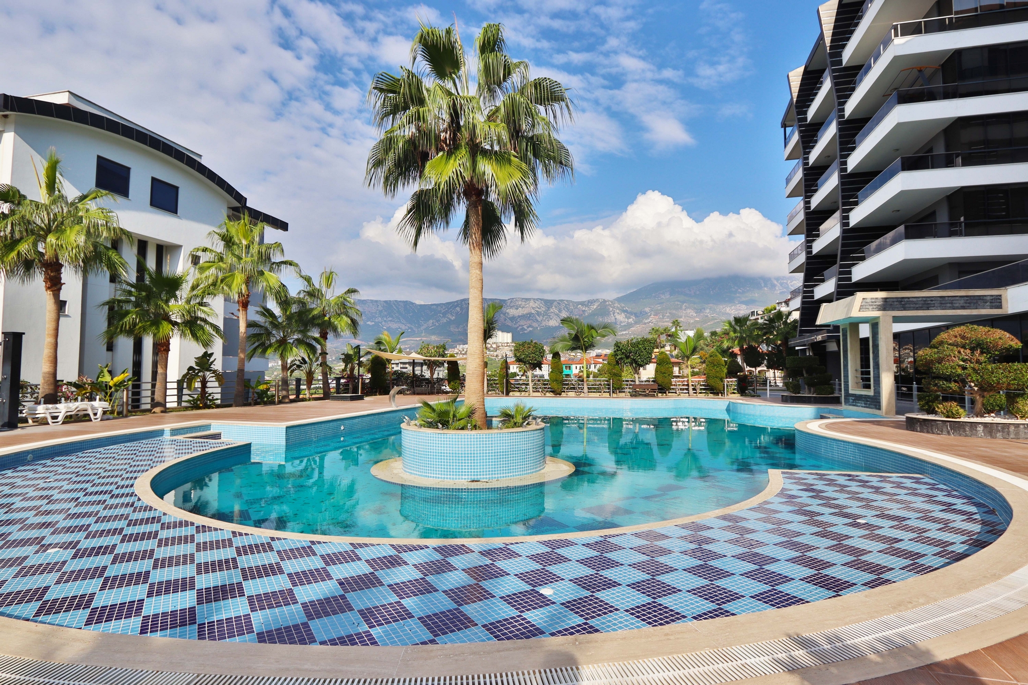 Round outdoor pool with a palm island, loungers and mountain views in a complex with flats in Kargicak
