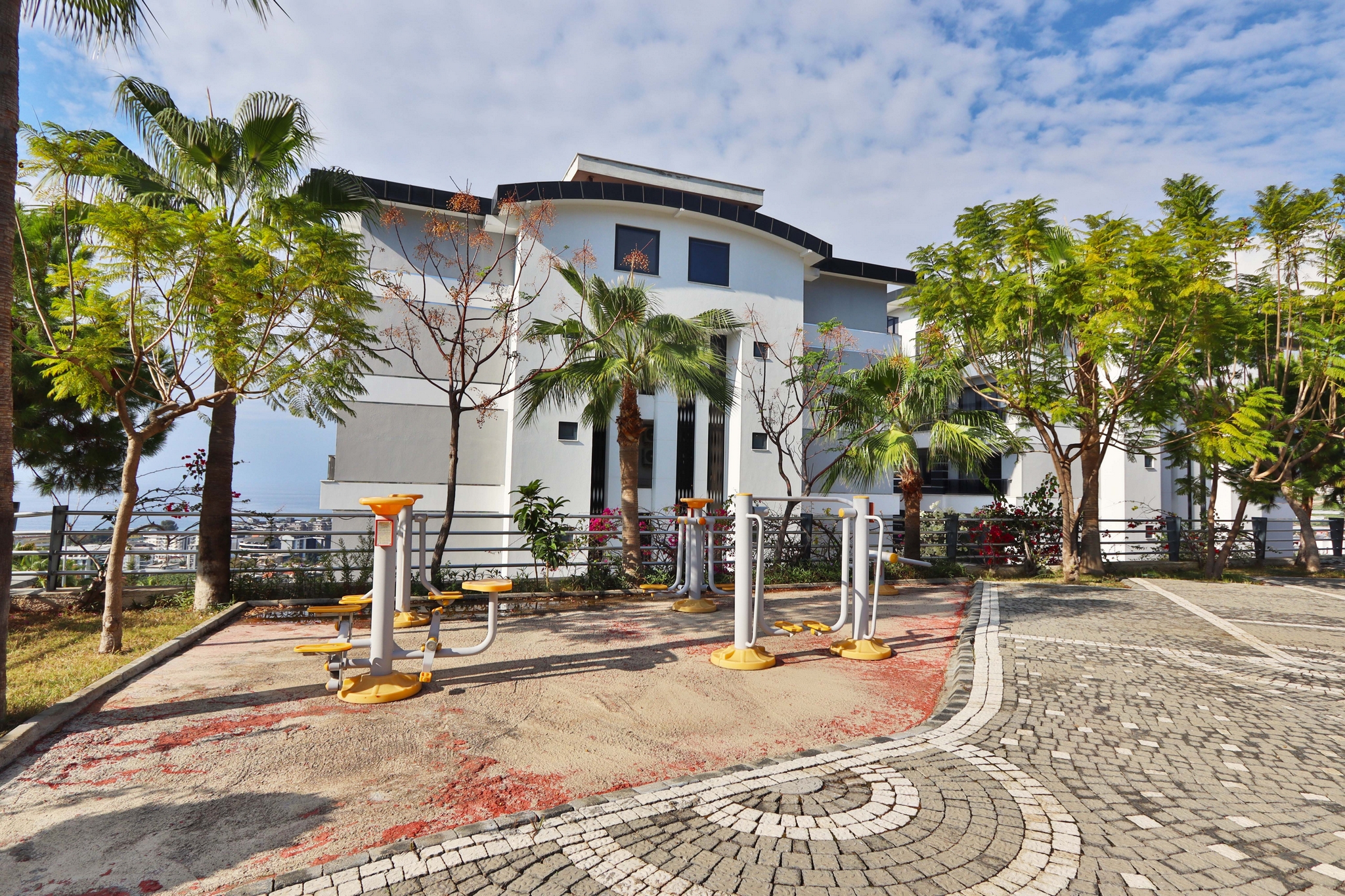 Outdoor fitness equipment beside a circular-patterned path and palms on a residence site in Kargicak, Turkey