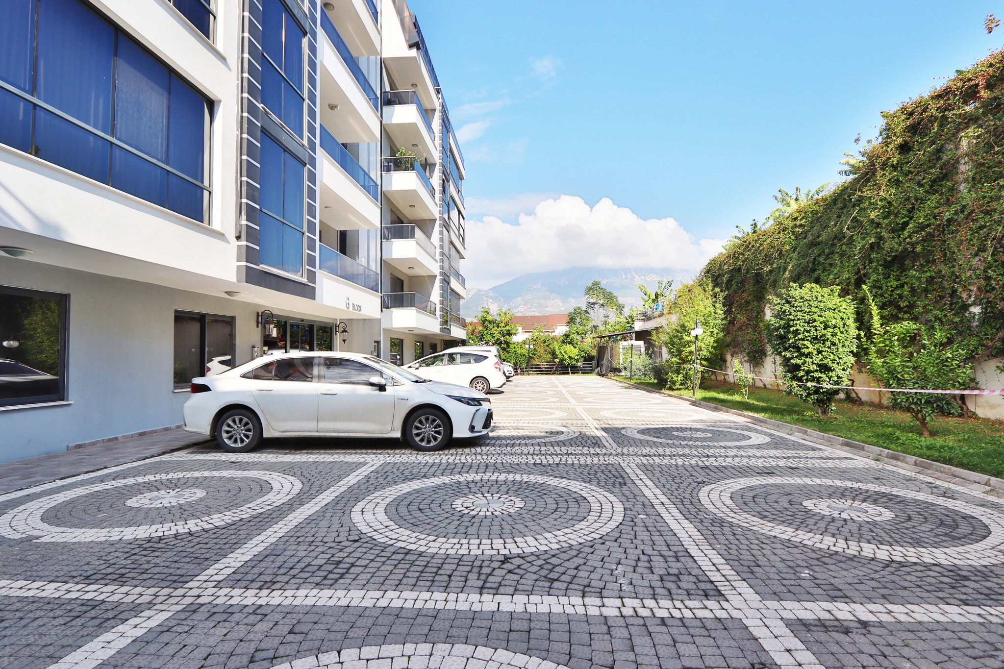 Open parking area with a car, hedges and a mountain slope beside apartments in Kargicak