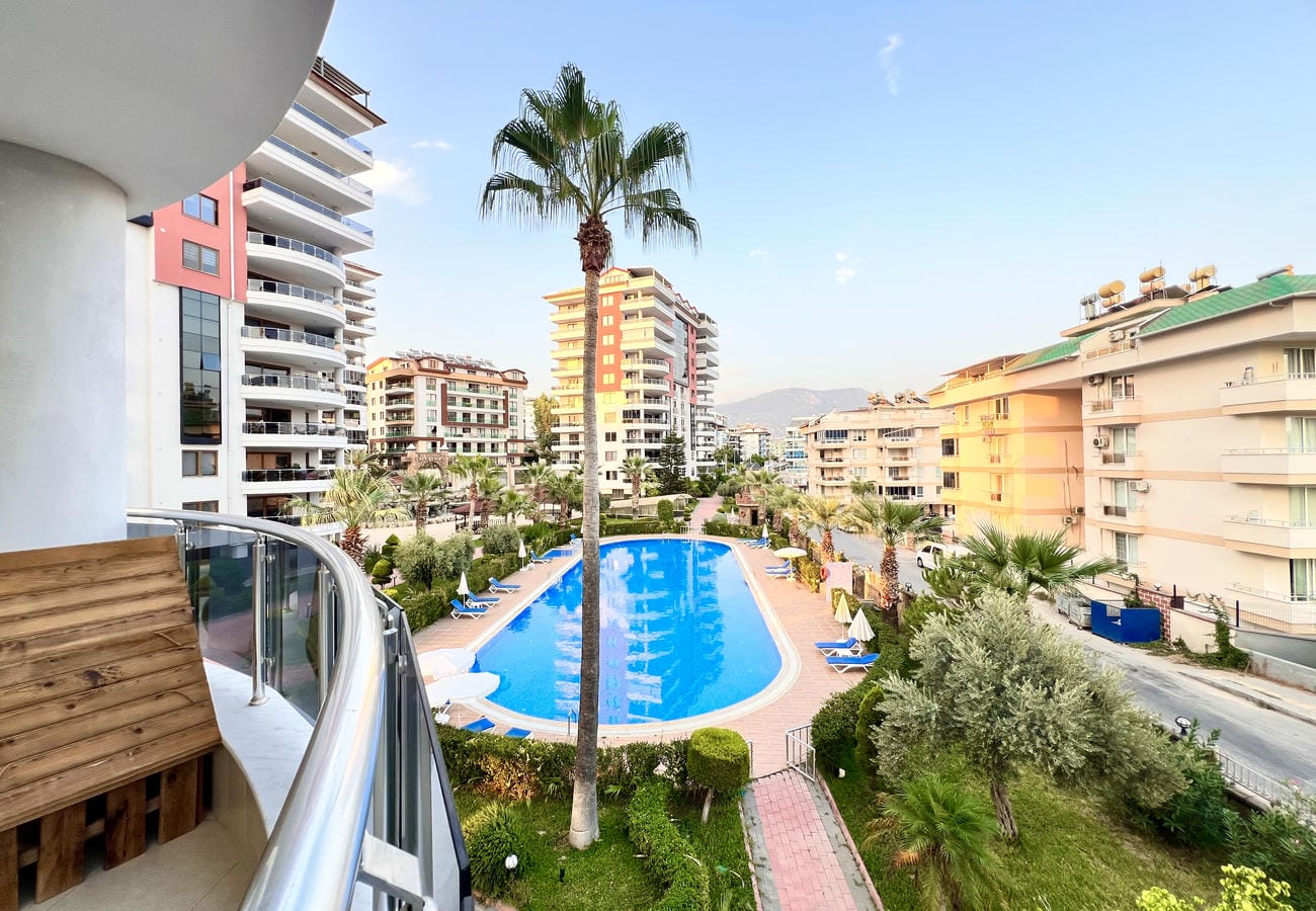 Cam korkuluklu balkon, satılık dairenin oval havuz ve dağ silüeti manzarasına açılıyor