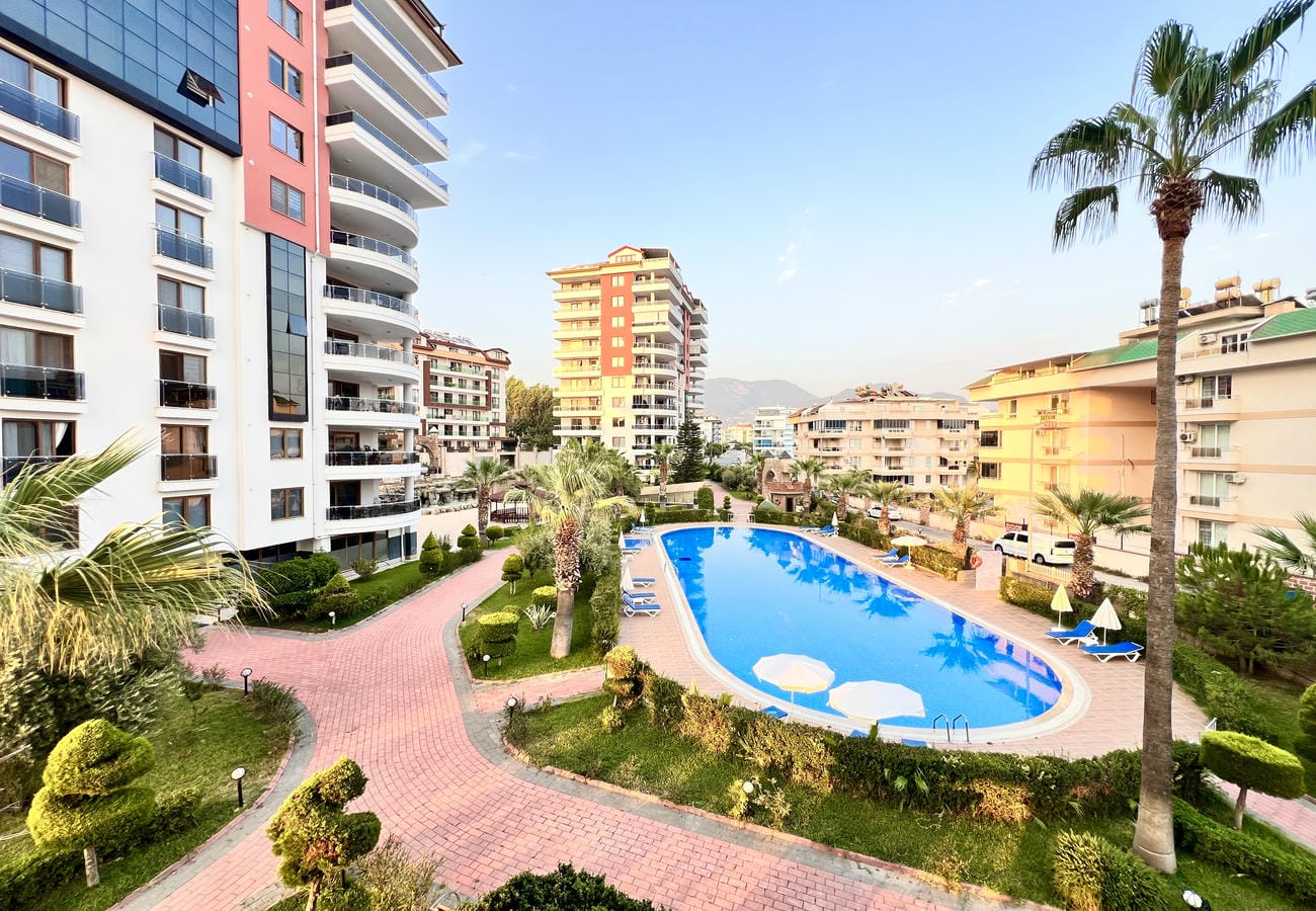 Palmiyeli oval havuz Türkiye’deki satıştaki rezidans alanında şezlonglar ve dağ silüetiyle görülüyor