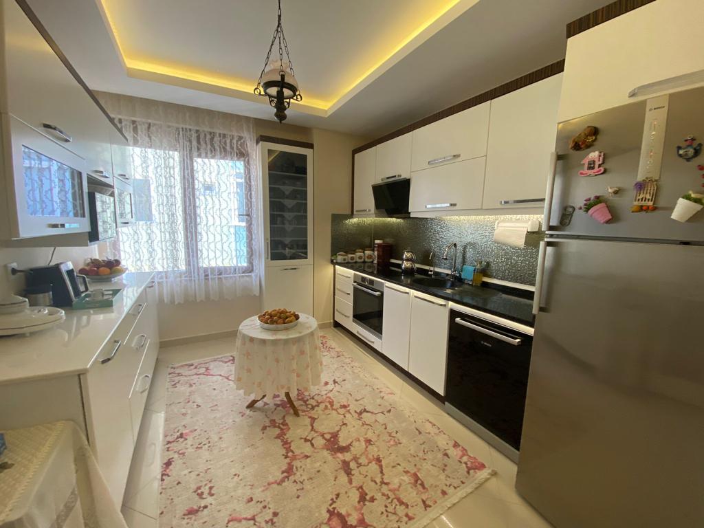 Siyah tezgâhlı beyaz mutfakta Alanya’daki çatı katı dairenin gümüş buzdolabı ve vitrin dolabı