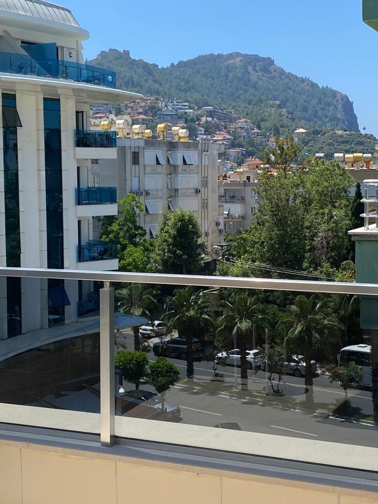 Cam korkuluklu balkondan Türkiye’deki rezidansın palmiyeli sokağı ve dağ yamacı görülüyor