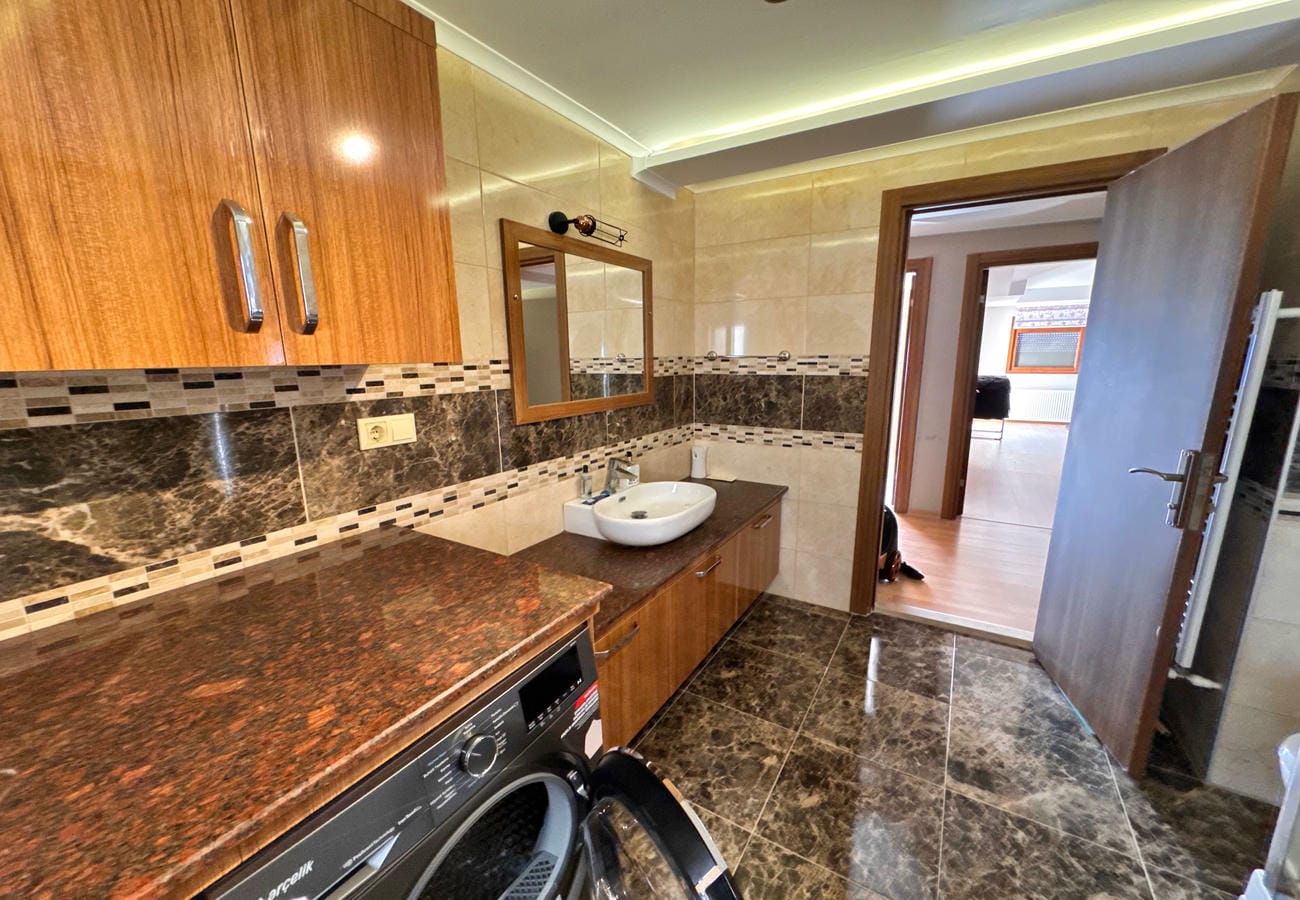 Banyoda ahşap dolaplı oval lavabo, çamaşır makinesi ve koyu mermer zemin