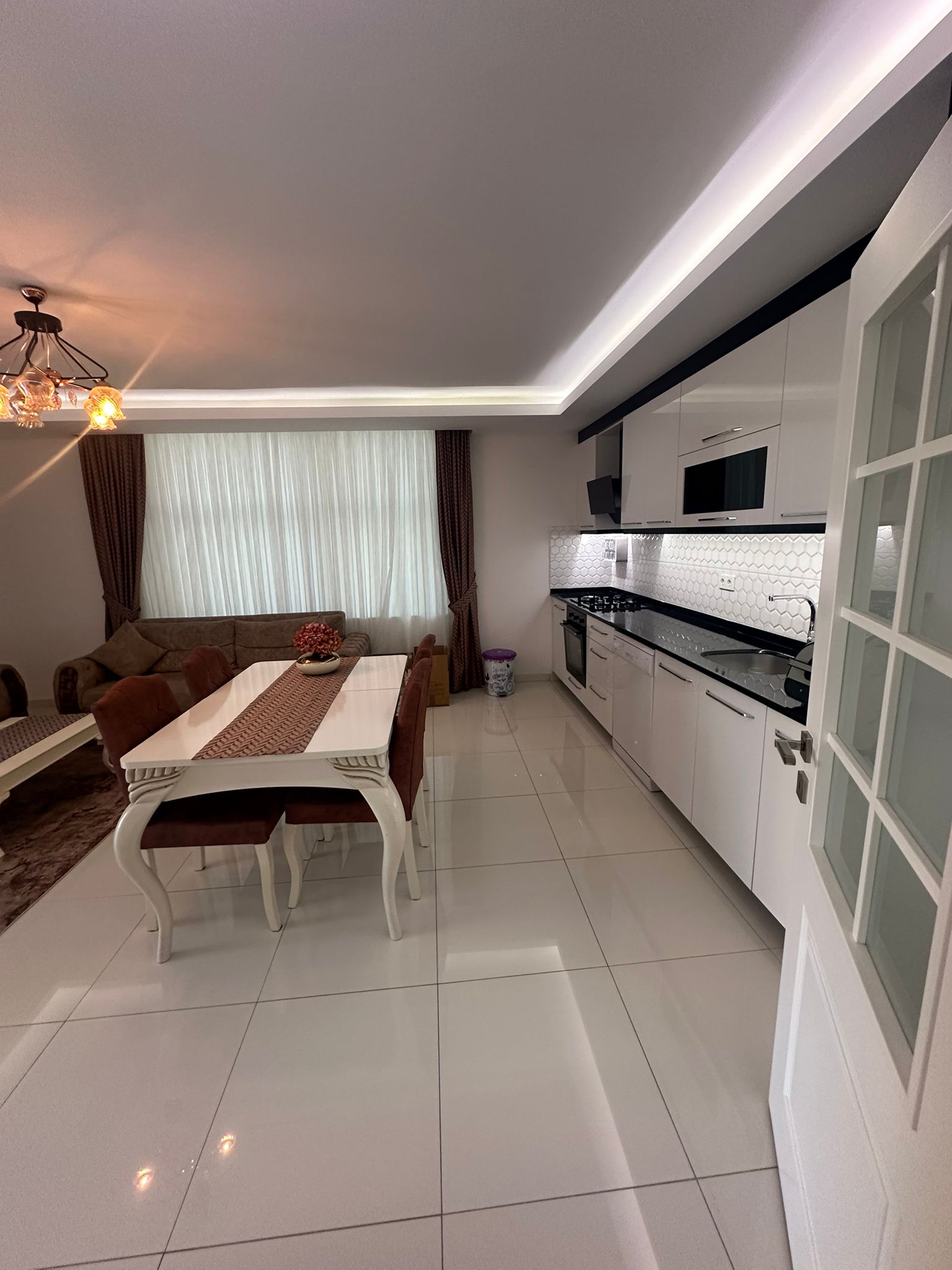 Dairenin mutfaklı salonunda tavana kadar beyaz dolaplar, siyah tezgâh ve yemek masası