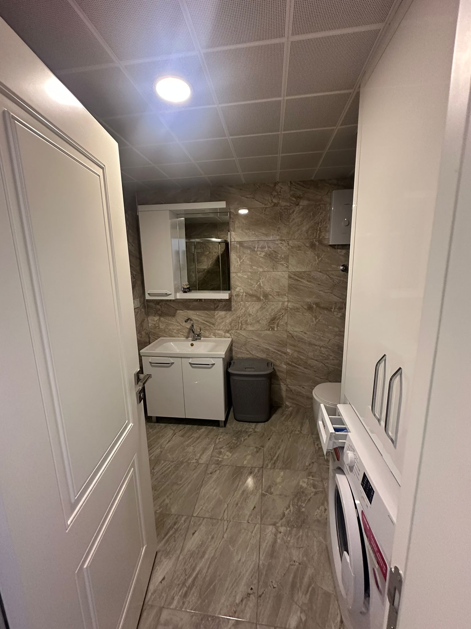 Dairenin banyosunda niş içindeki çamaşır makinesi, yüksek dolap ve aynalı lavabo ünitesi